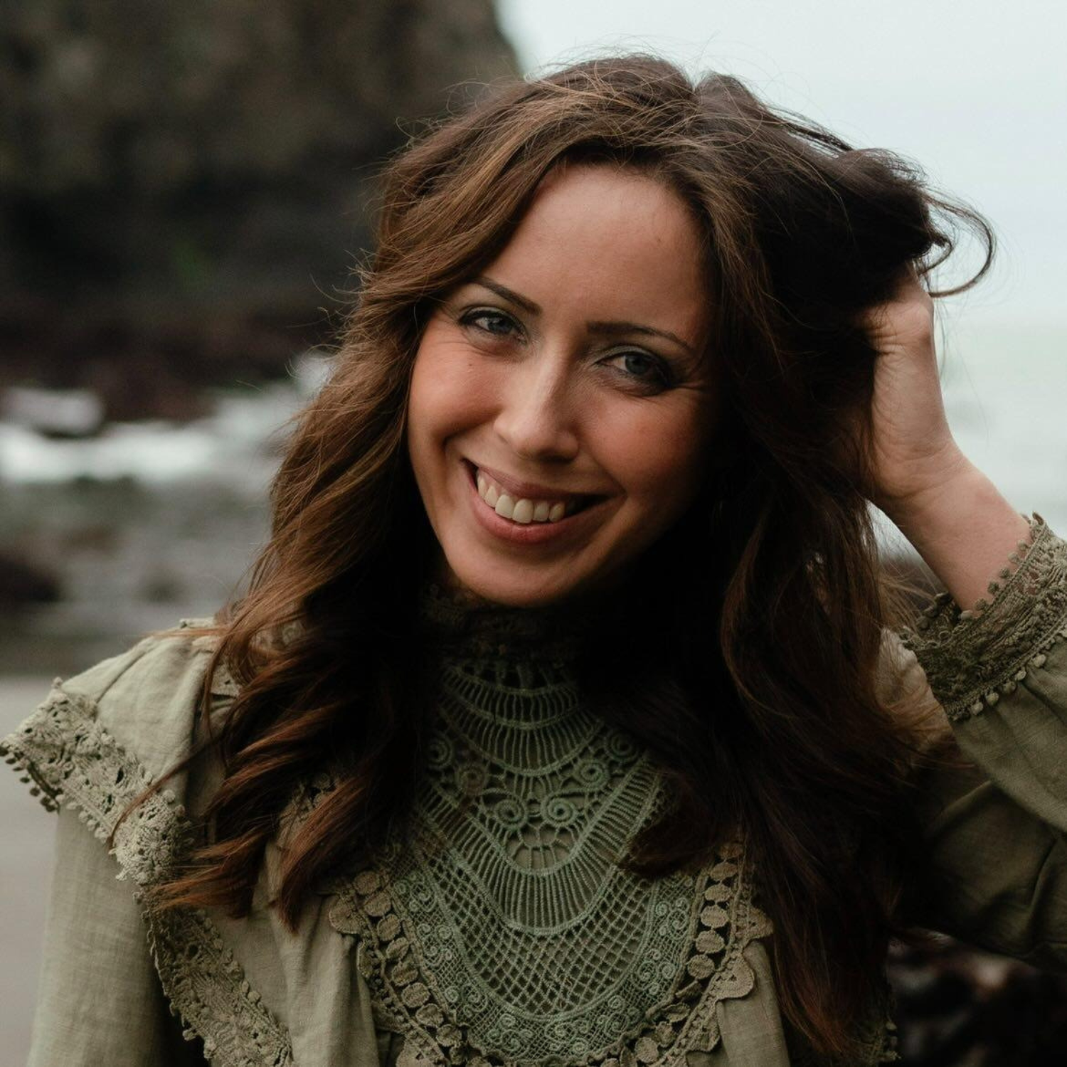 Smiling woman outdoors with wavy brown hair, wearing a green lace top, standing by a rocky coastline.
