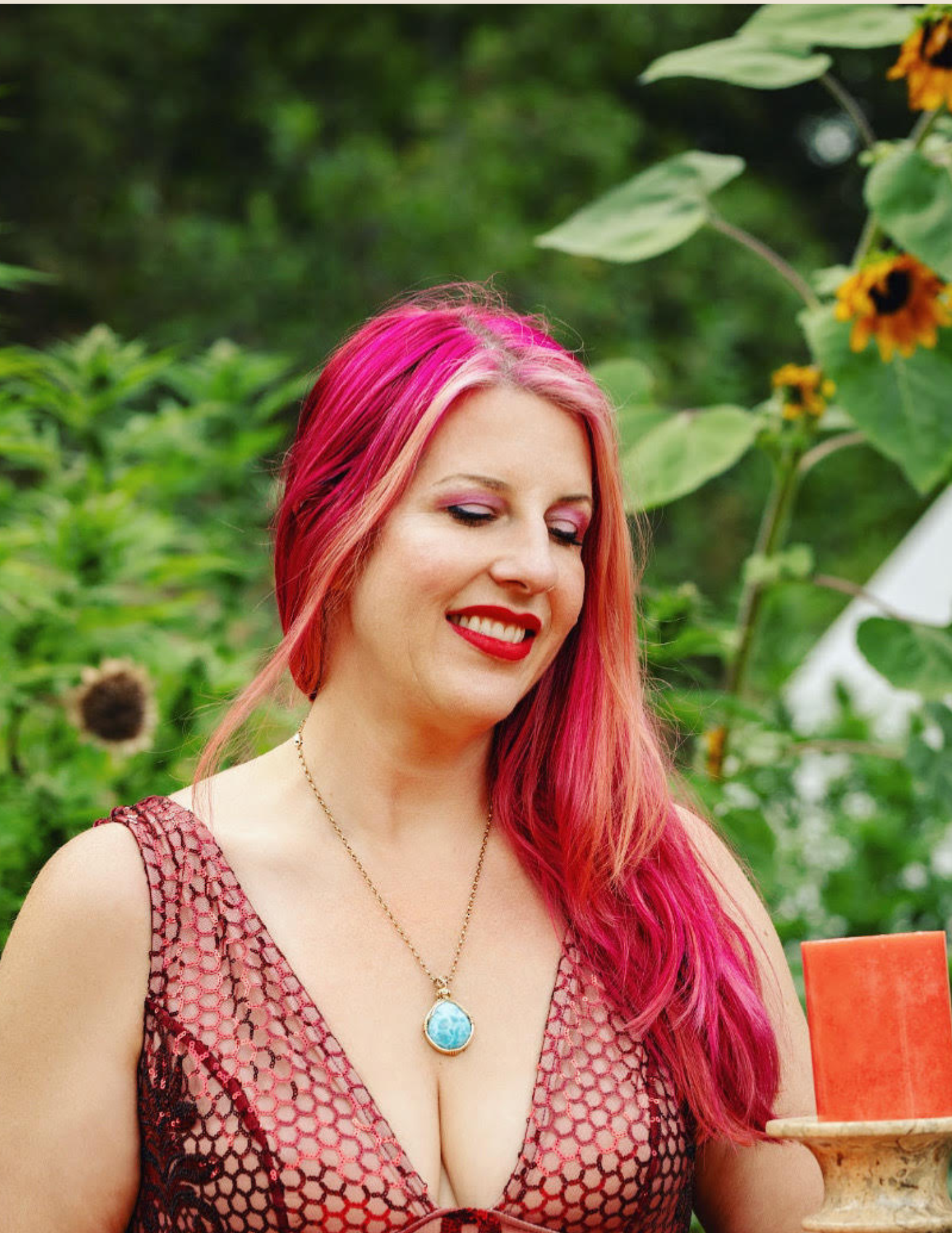 Woman with pink hair and a patterned dress outdoors near sunflowers.