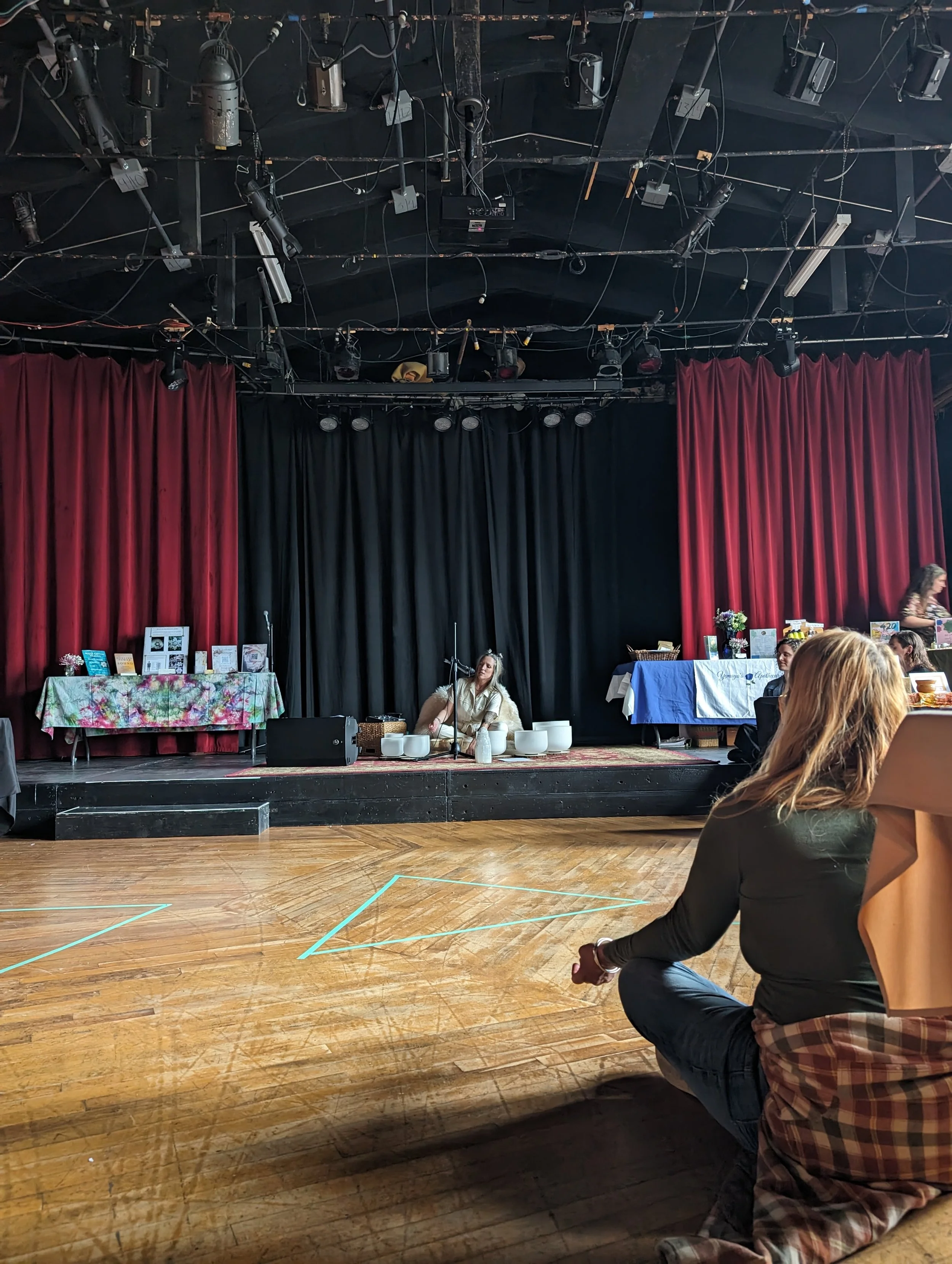Interior of a dimly lit venue with a stage featuring a person sitting on the floor surrounded by white bowls and a microphone. Red and black curtains are in the background. A person is sitting on the wooden floor in the foreground, facing the stage.