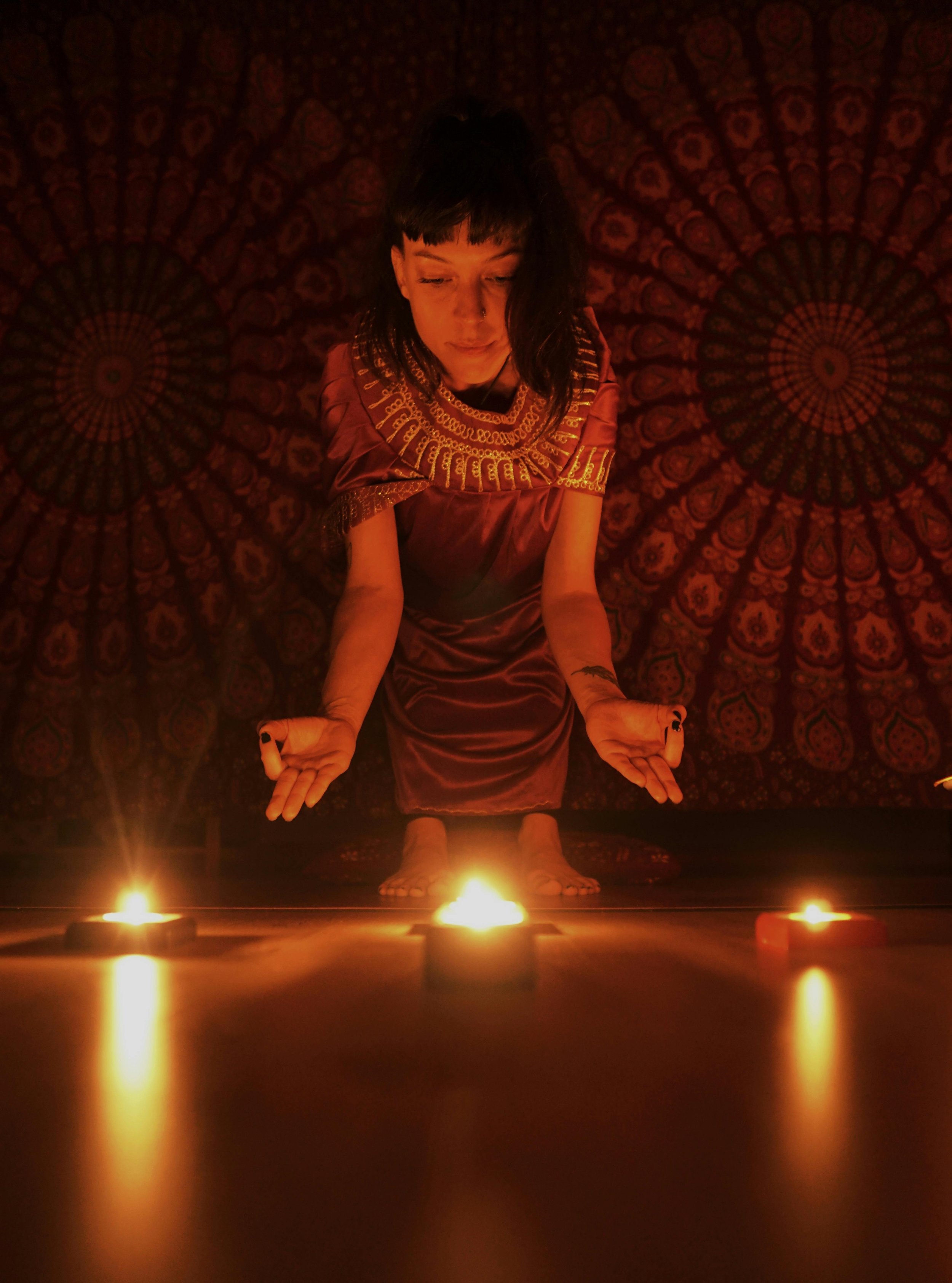 Person in dim light performing a ritual with candles on the floor, dressed in a red and gold robe, with a patterned tapestry background.