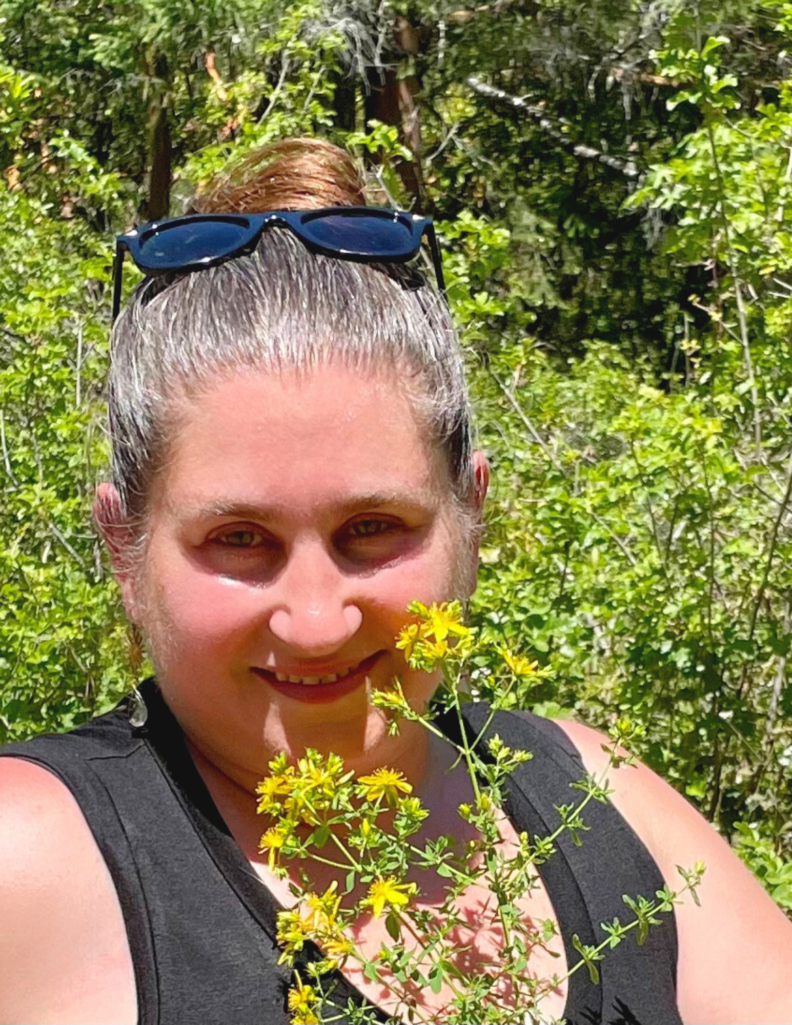 Person with sunglasses on head, smiling and holding a bunch of yellow wildflowers, surrounded by greenery.