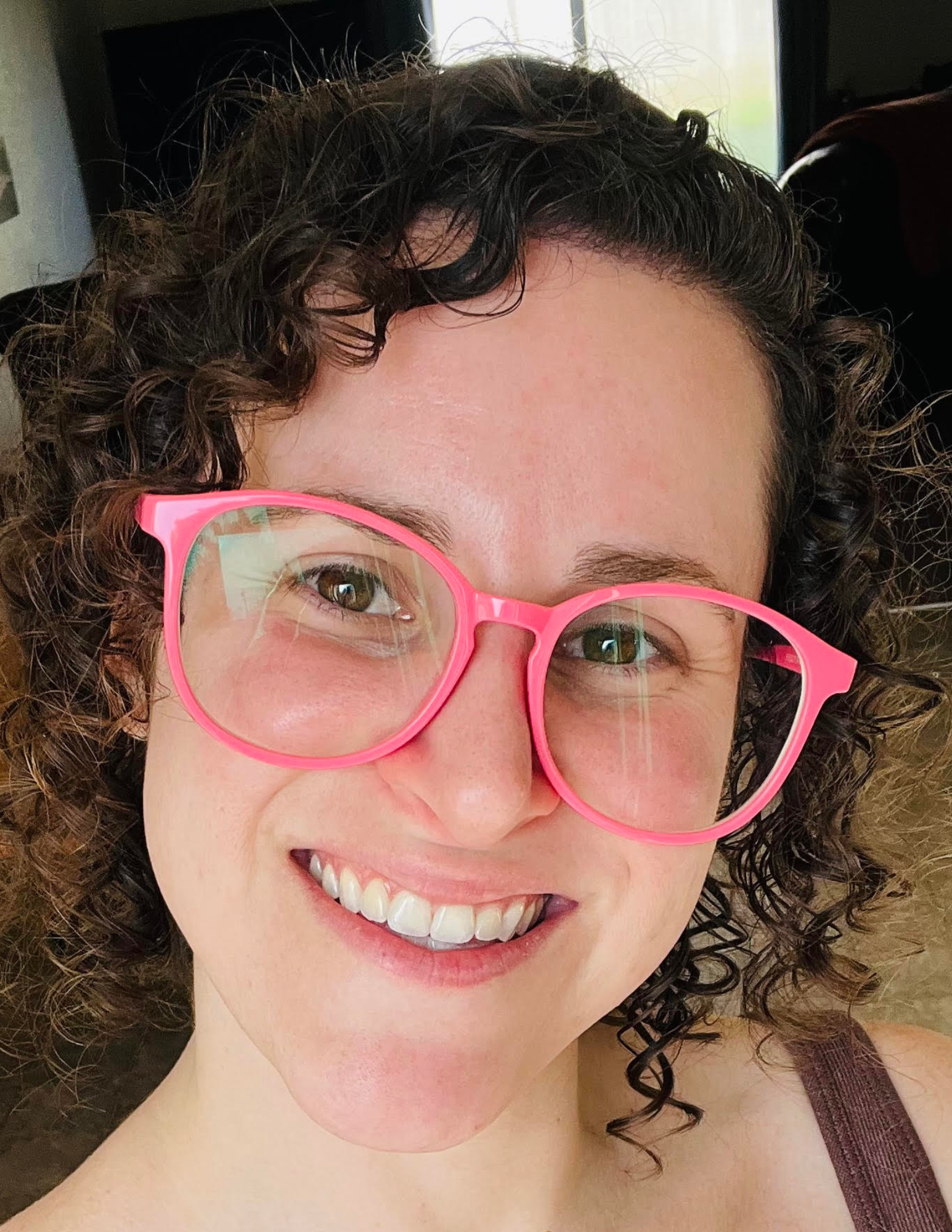 Person smiling wearing large pink glasses with curly hair, indoors.