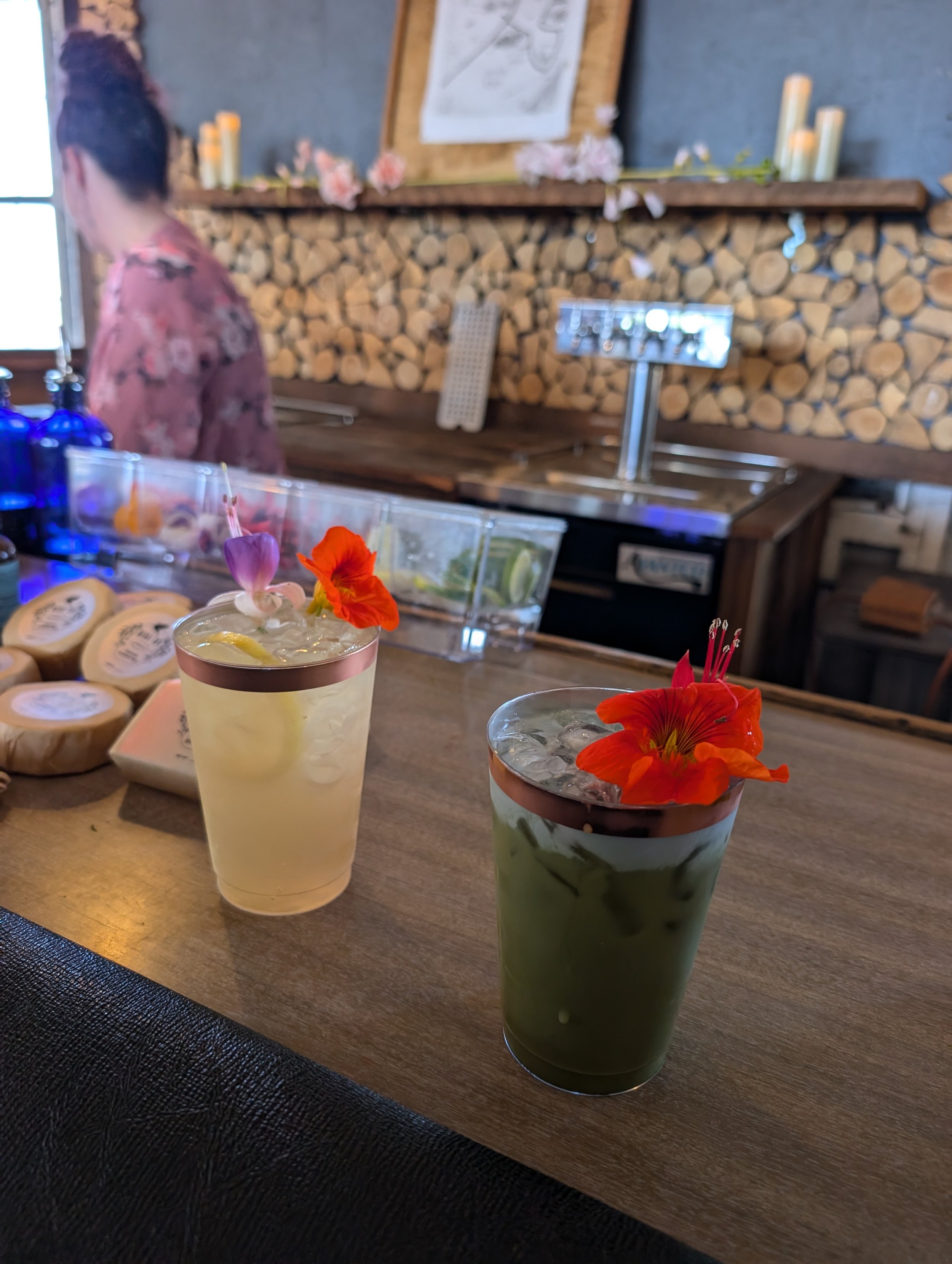 Two beverages on a wooden counter, one green with an orange flower and the other light yellow with a lemon slice and a purple flower, in a cafe with a patterned wooden wall background.