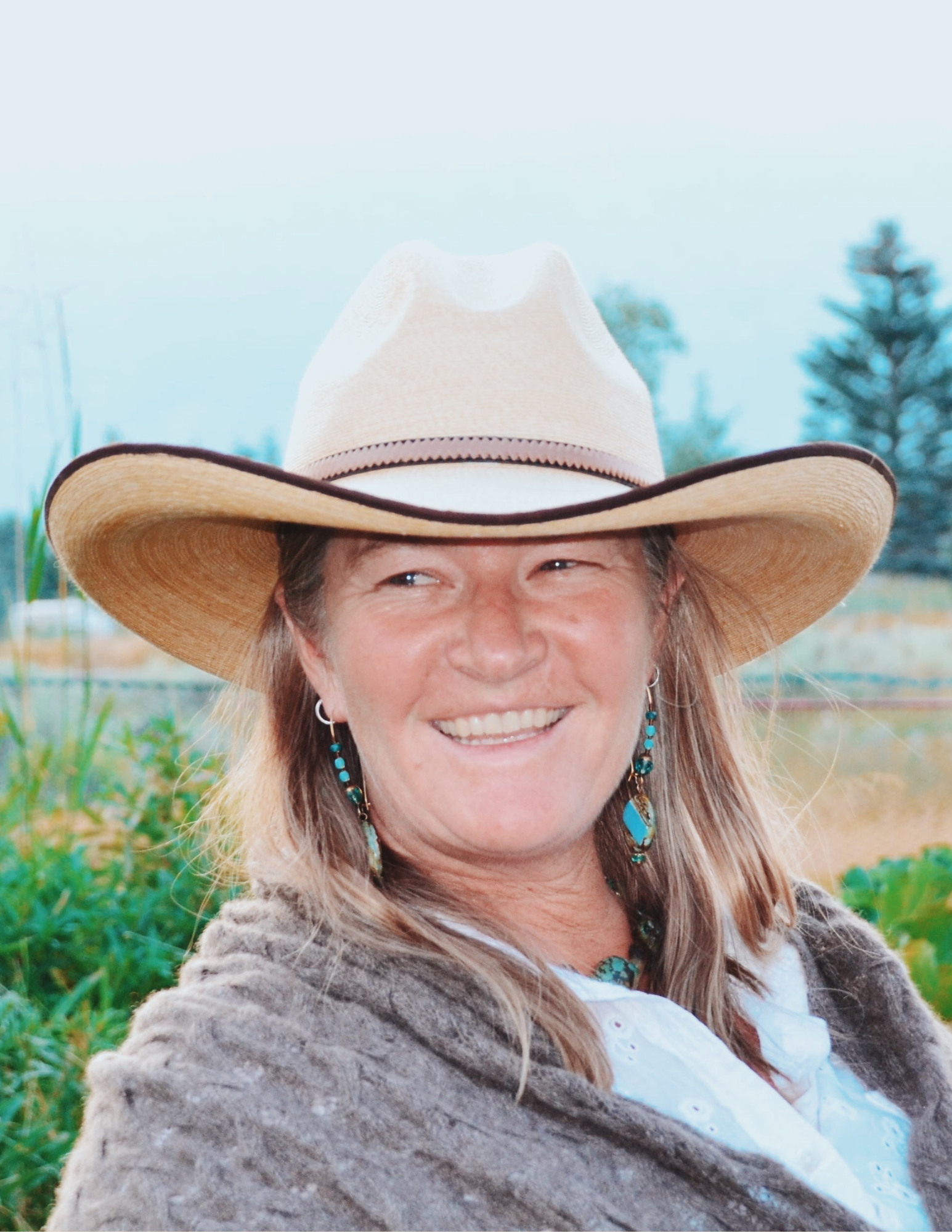 Person wearing a cowboy hat and earrings, smiling, with a natural outdoor background.