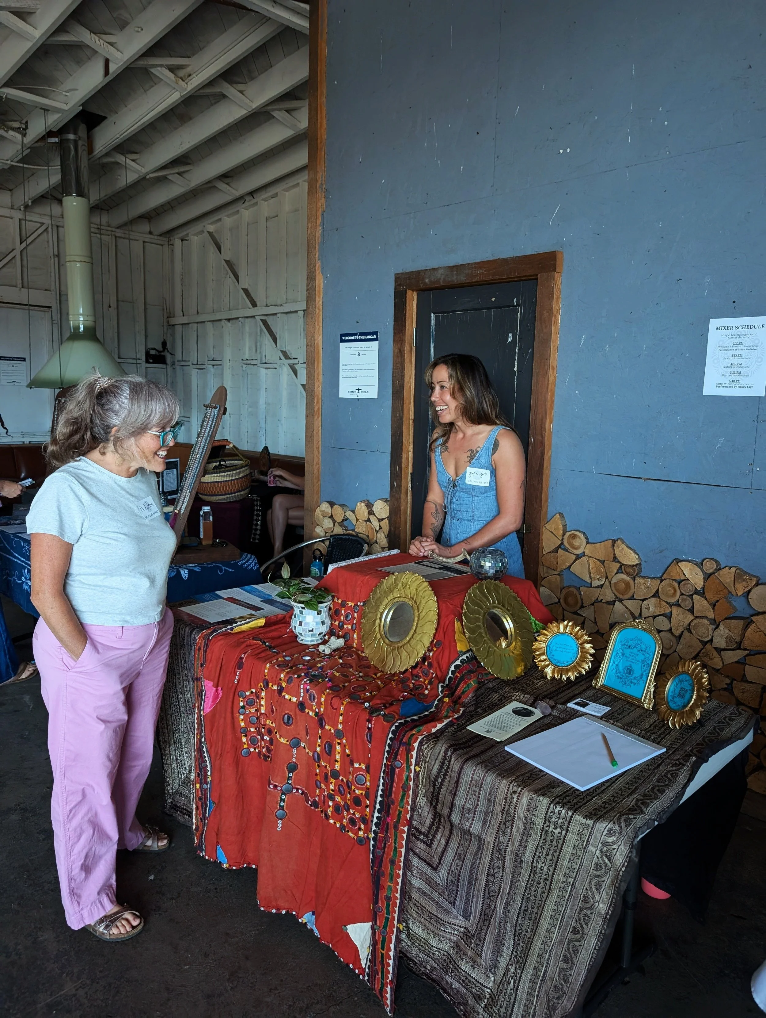 Two women interacting at a decorated table inside a rustic indoor space. The table is draped with colorful textiles and adorned with framed items and decorative objects. Firewood is stacked against the wall nearby.