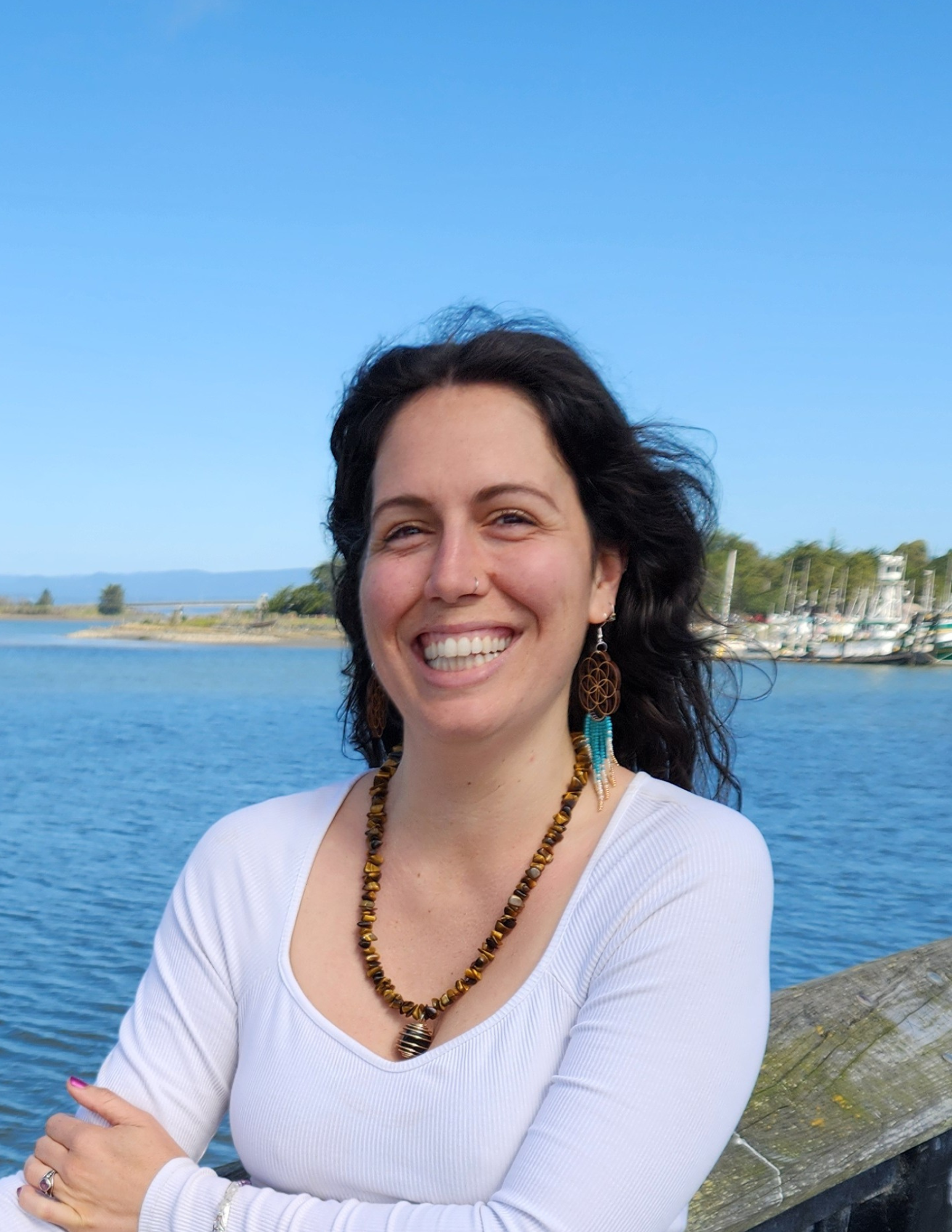 Woman smiling near a body of water with sailboats in the background, wearing a white top and beaded necklace.