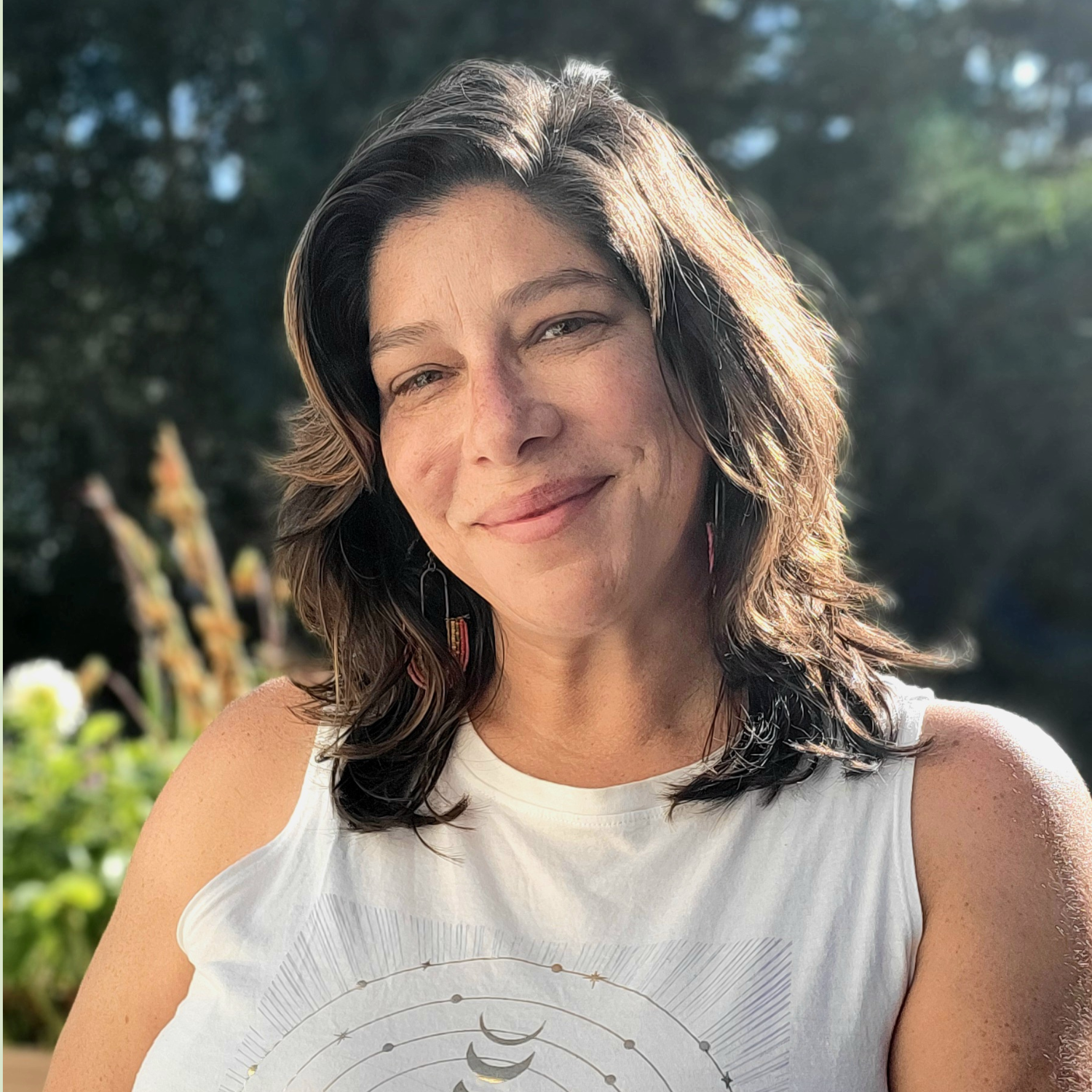 A smiling woman with brown hair wearing a white tank top featuring a celestial design, standing outdoors with greenery in the background.