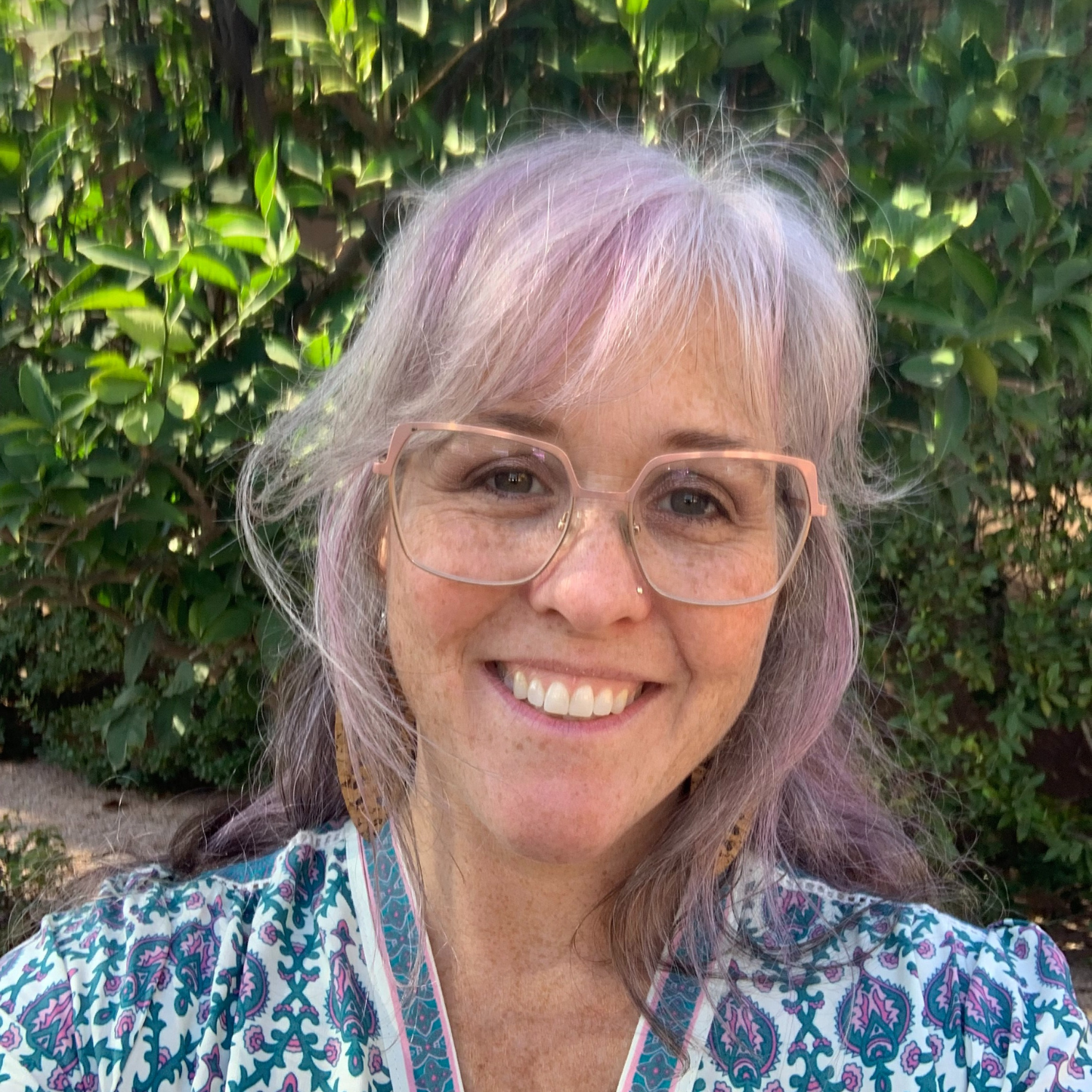 Smiling woman with glasses and purple-tinted hair standing outdoors, surrounded by greenery.