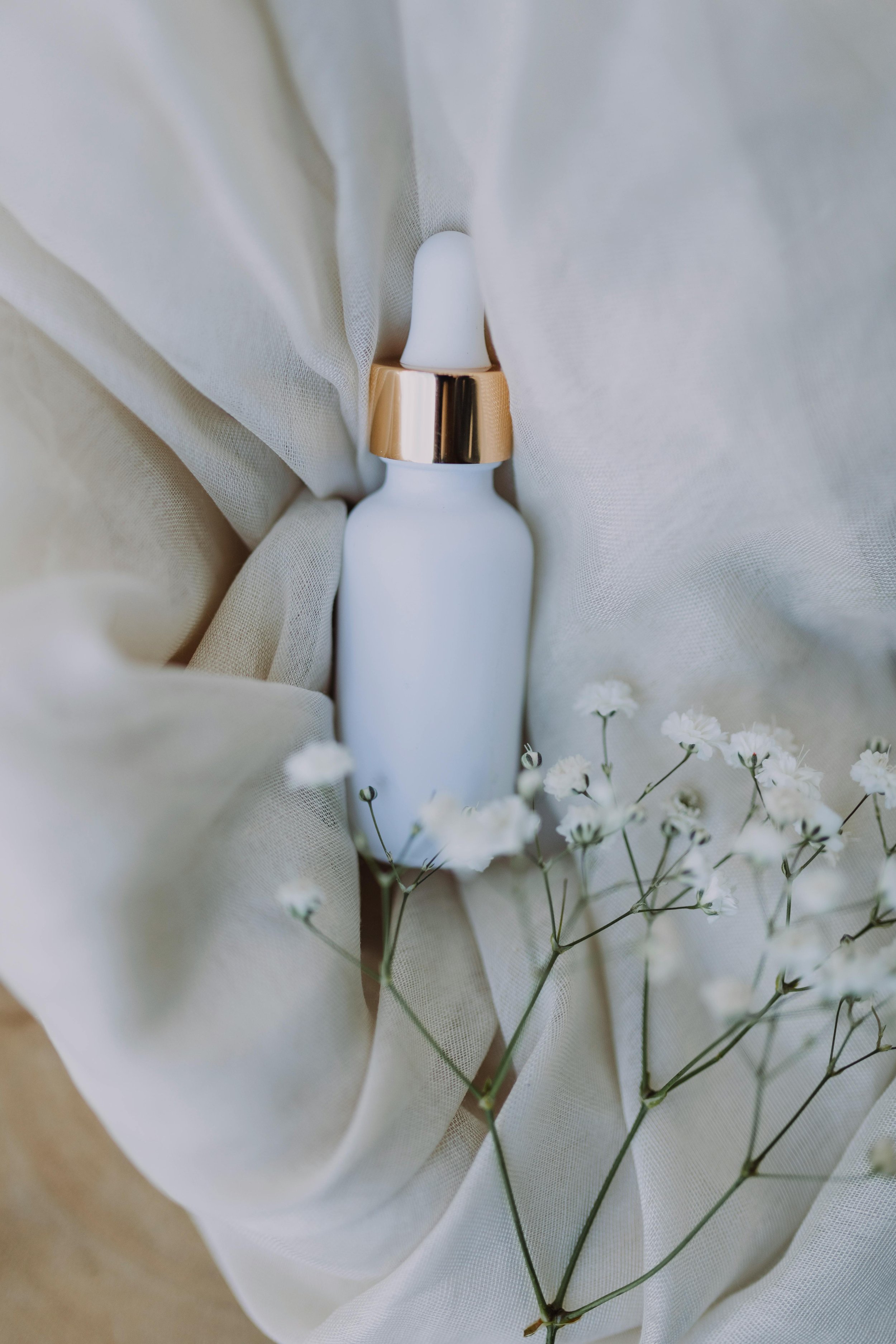 White dropper bottle with gold cap on soft fabric and small white flowers