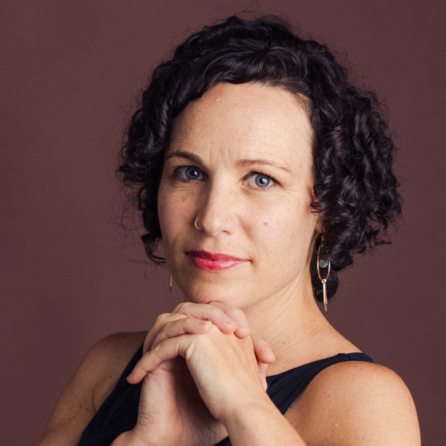 Portrait of a person with curly dark hair and a nose piercing, wearing earrings and a sleeveless top, against a maroon background.