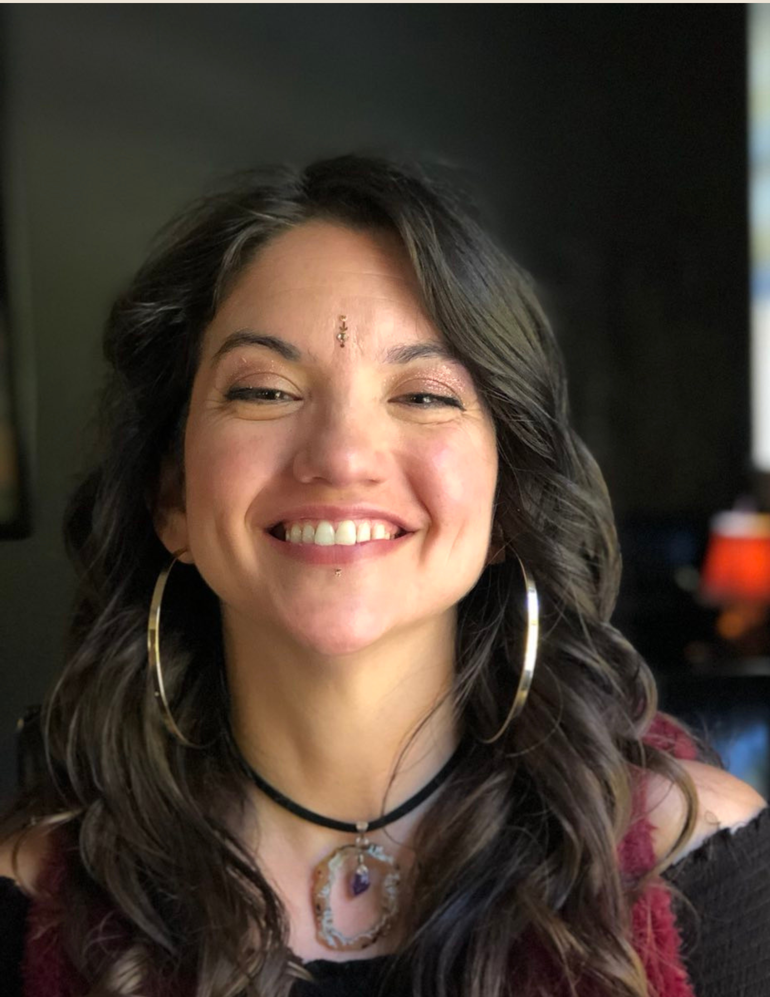 Smiling person with long dark hair, wearing large hoop earrings and gemstone necklace, against a dark background.