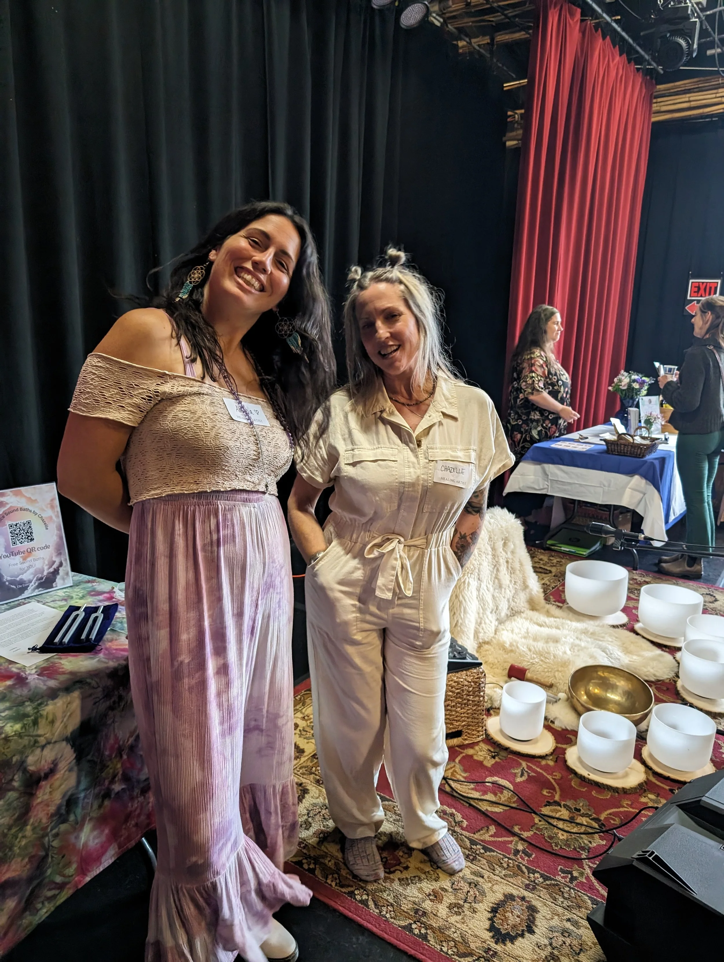 Two women smiling at an event with a spiritual or wellness theme. One is wearing a purple off-the-shoulder dress, and the other is in a white jumpsuit. They stand near a display with singing bowls and a rug. Other attendees and tables are visible in 
