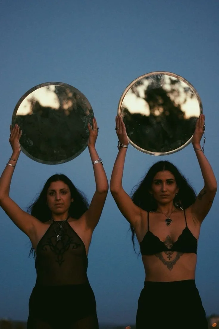 Two women holding circular mirrors above their heads against a twilight sky.