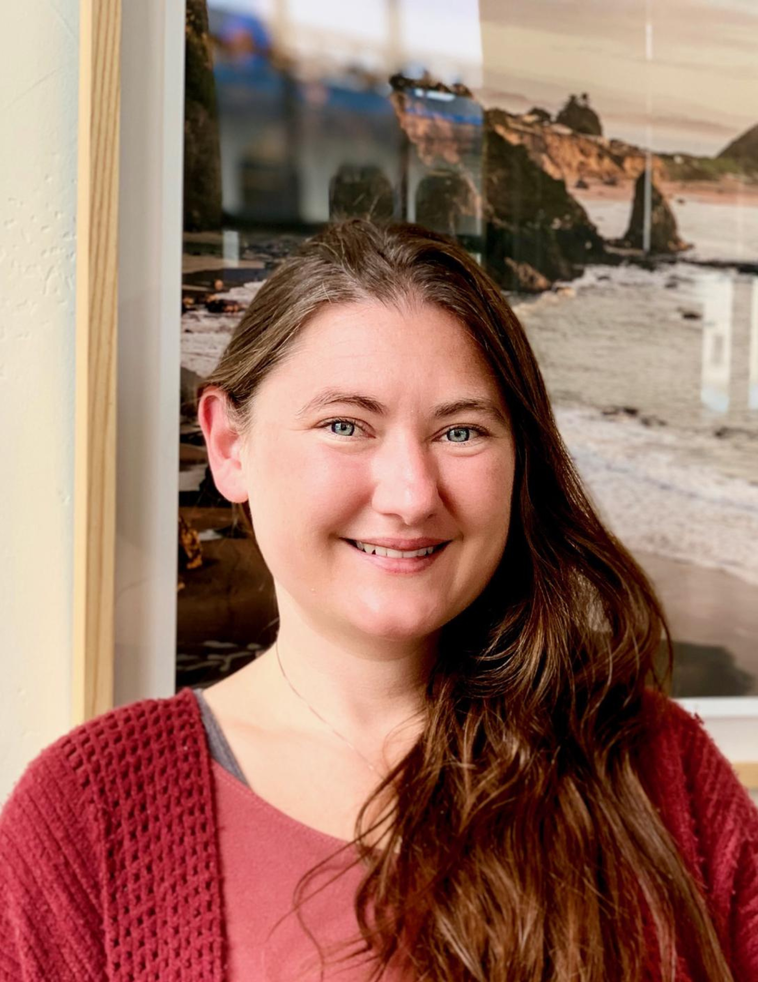 Person smiling indoors with long hair and wearing a red cardigan, standing in front of a framed landscape photograph of a rocky coastline.