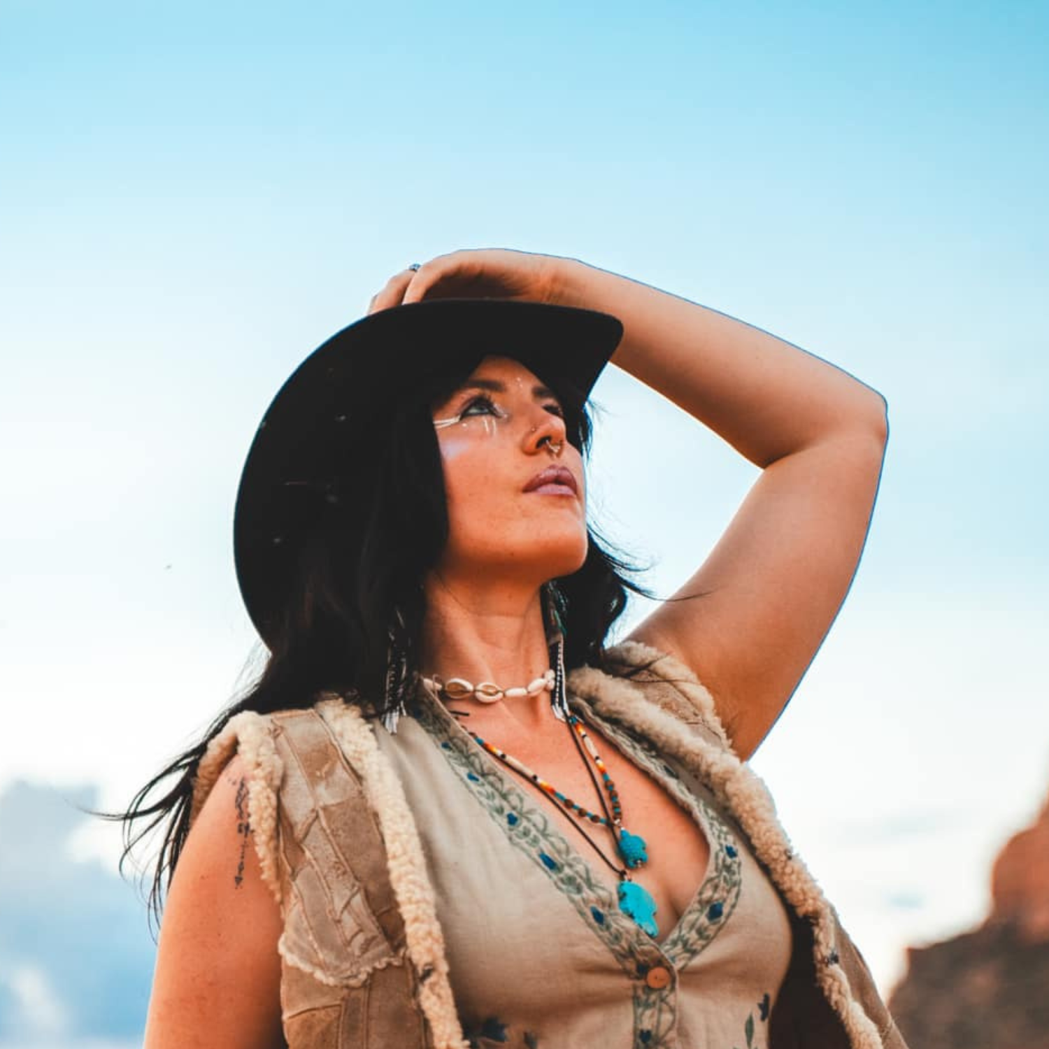 Woman wearing a cowboy hat and a beige vest, looking upwards against a blue sky.