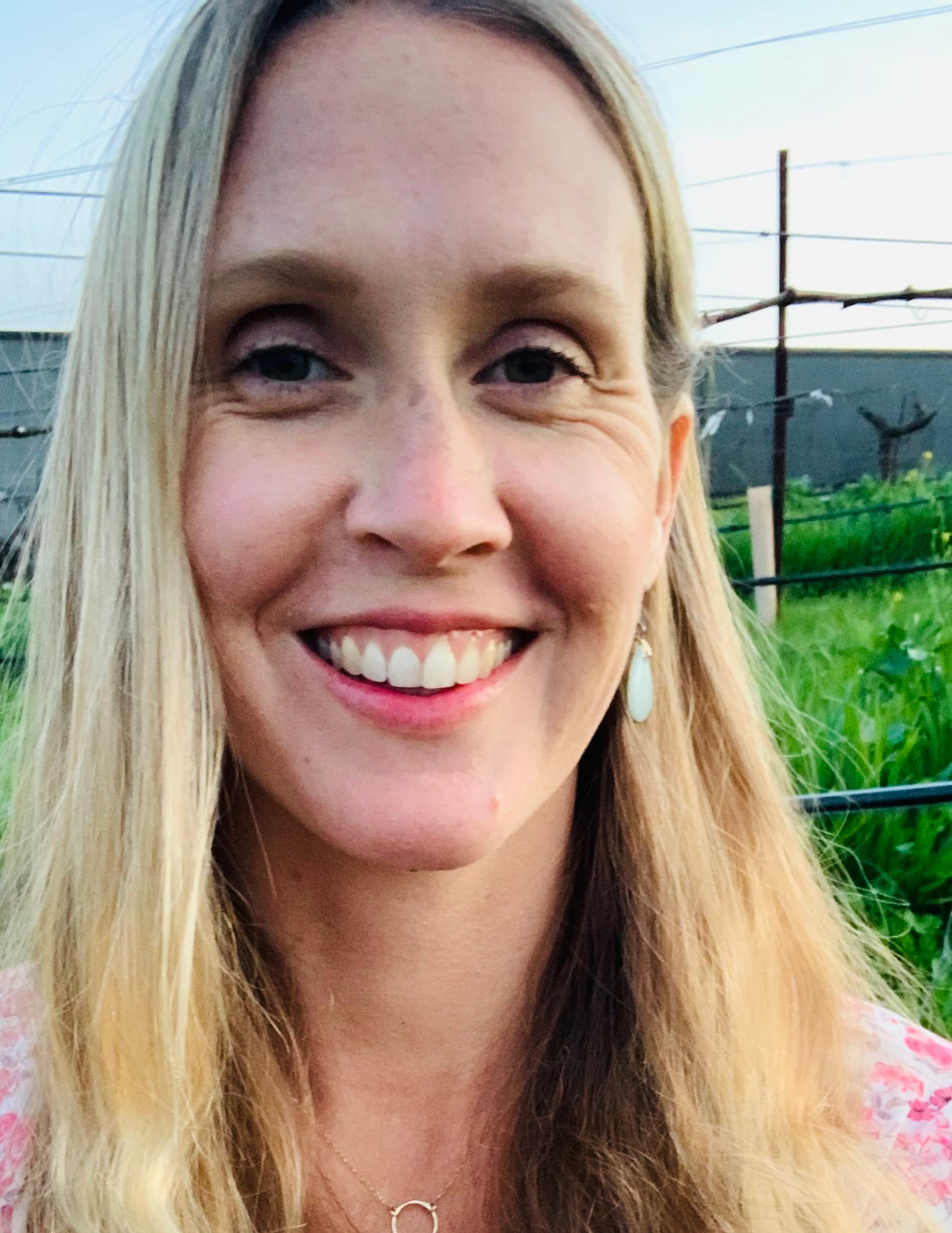 A woman smiling outdoors with green plants in the background.