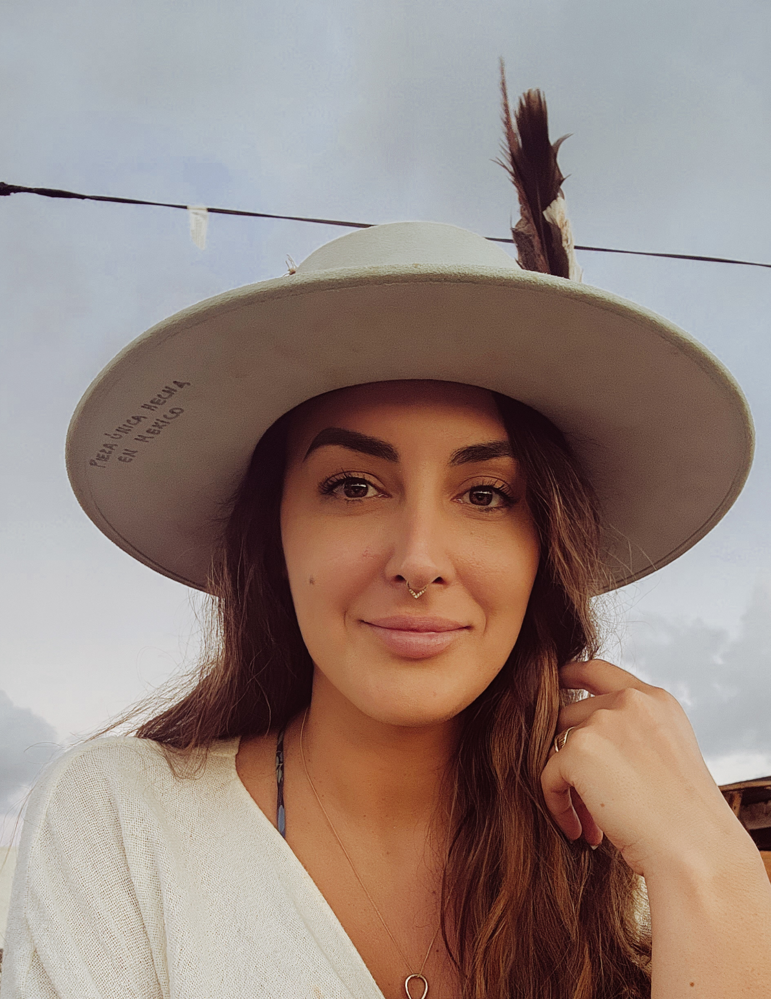 Person wearing a wide-brimmed hat with a feather and a septum ring, smiling outdoors.