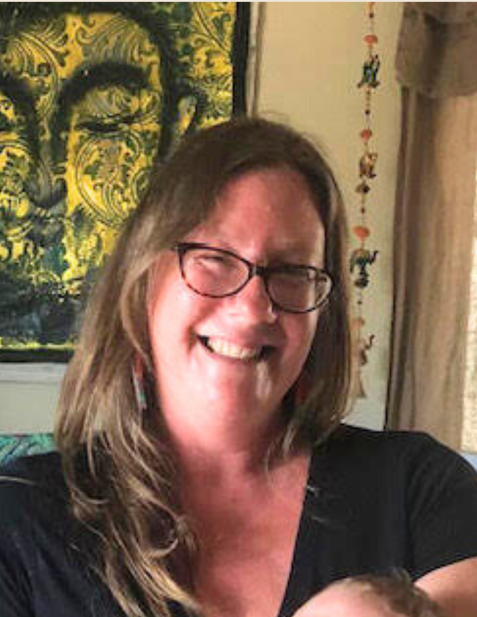 A smiling woman with glasses and long hair, sitting indoors. Behind her is a decorative wall hanging with a Buddha face design and a string of hanging ornaments.