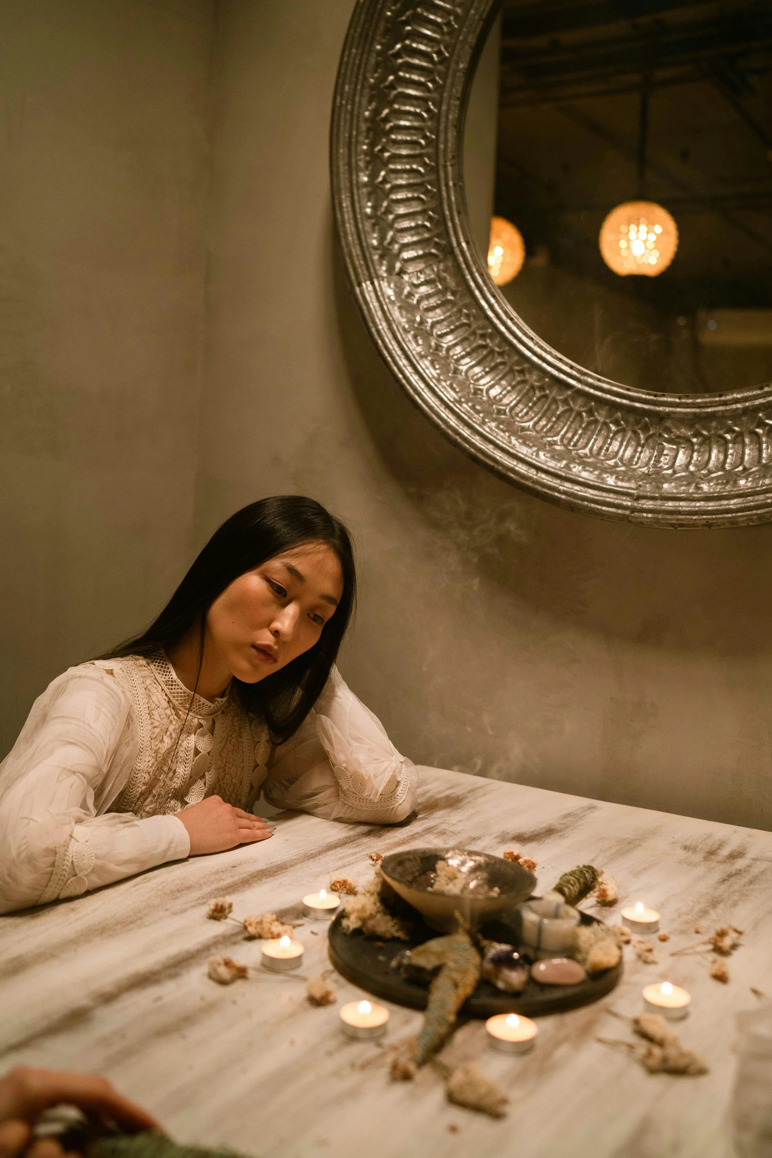 A person with long black hair in a white lace dress sitting at a table with lit candles and stones, looking contemplative. A round, ornate mirror is on the wall, reflecting warm lighting.