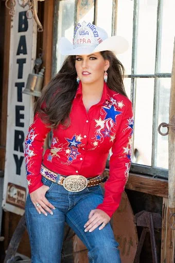 Woman wearing a white cowboy hat with 'Rodeo' and 'USA' embroidery, red western shirt with star and horse designs, blue jeans, and a large belt buckle, standing indoors in front of a window with rustic decor.