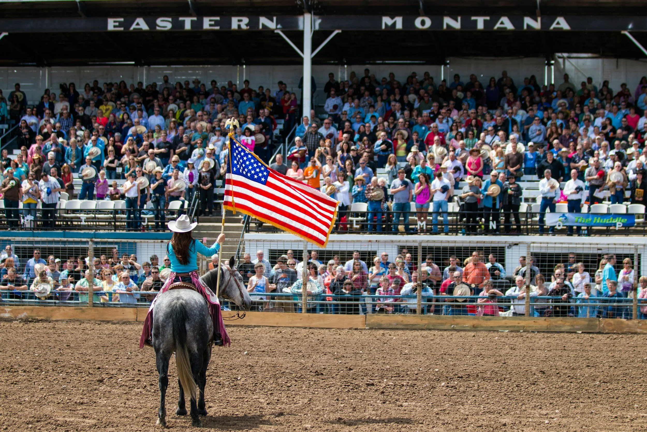 A rodeo scene with a woman on a horse holding an American flag, facing a crowd in the stands at the Eastern Montana rodeo event.