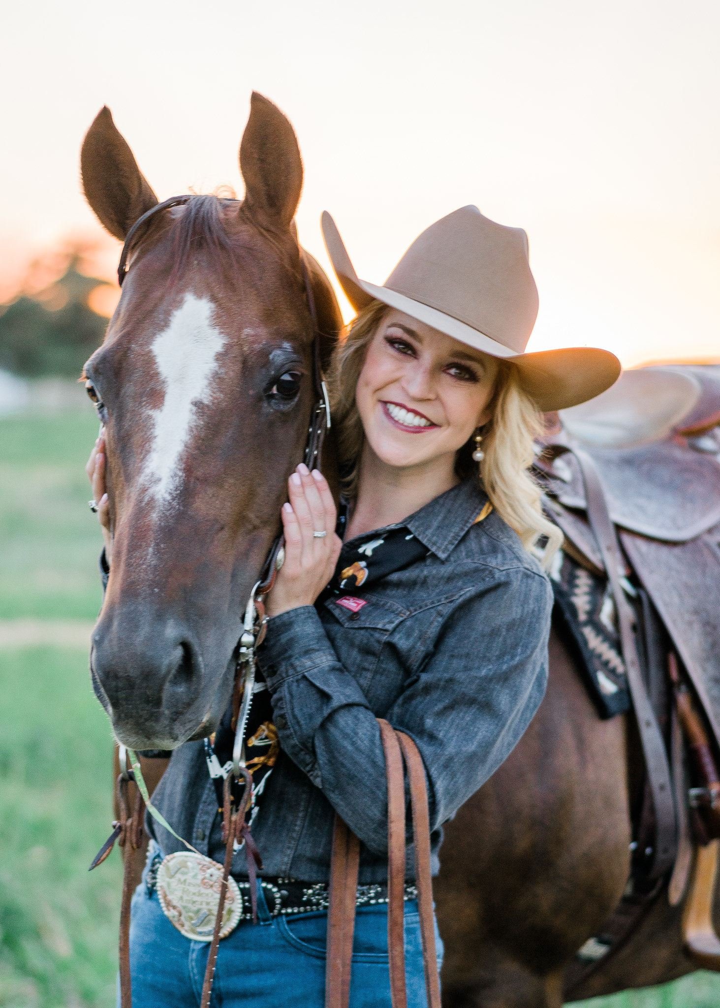 A woman smiling and hugging a brown horse with a white stripe on its face, outdoors during sunset.