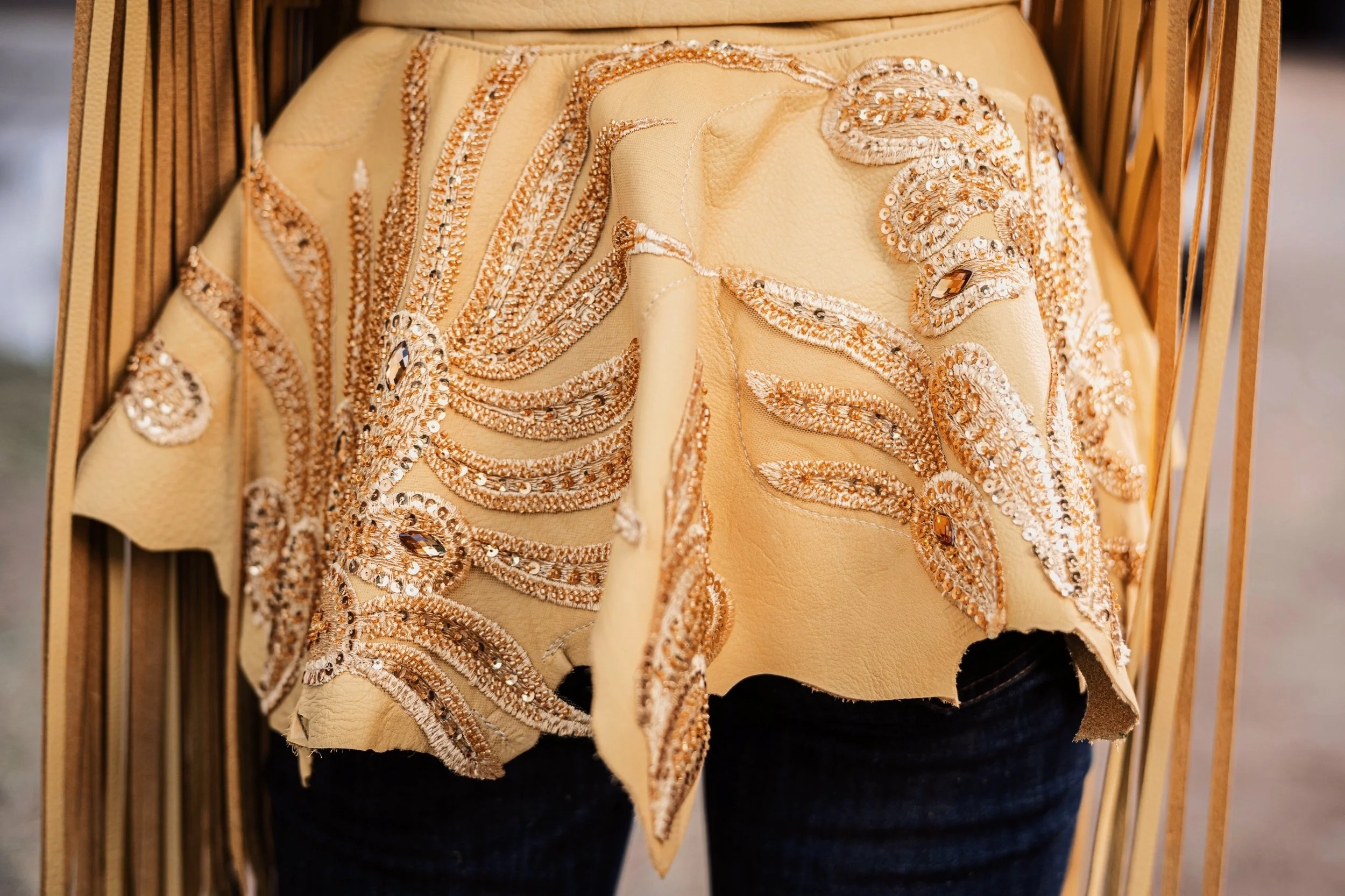 Close-up of a beige leather jacket with intricate bead and sequin embroidery in floral and swirl patterns, hanging on a wooden rack.