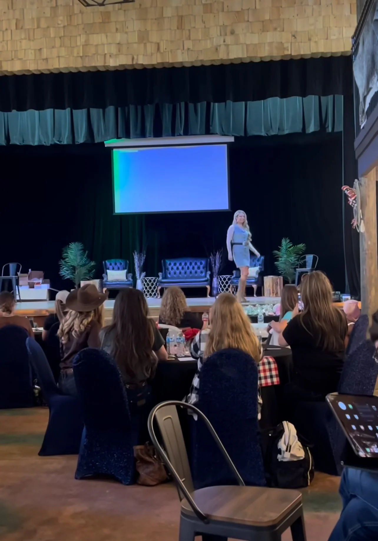 A woman standing on a stage in front of an audience in a large hall with wooden walls. The stage is decorated with plants and furniture, including chairs and a sofa. There is a large screen behind the woman displaying a blue and green gradient.