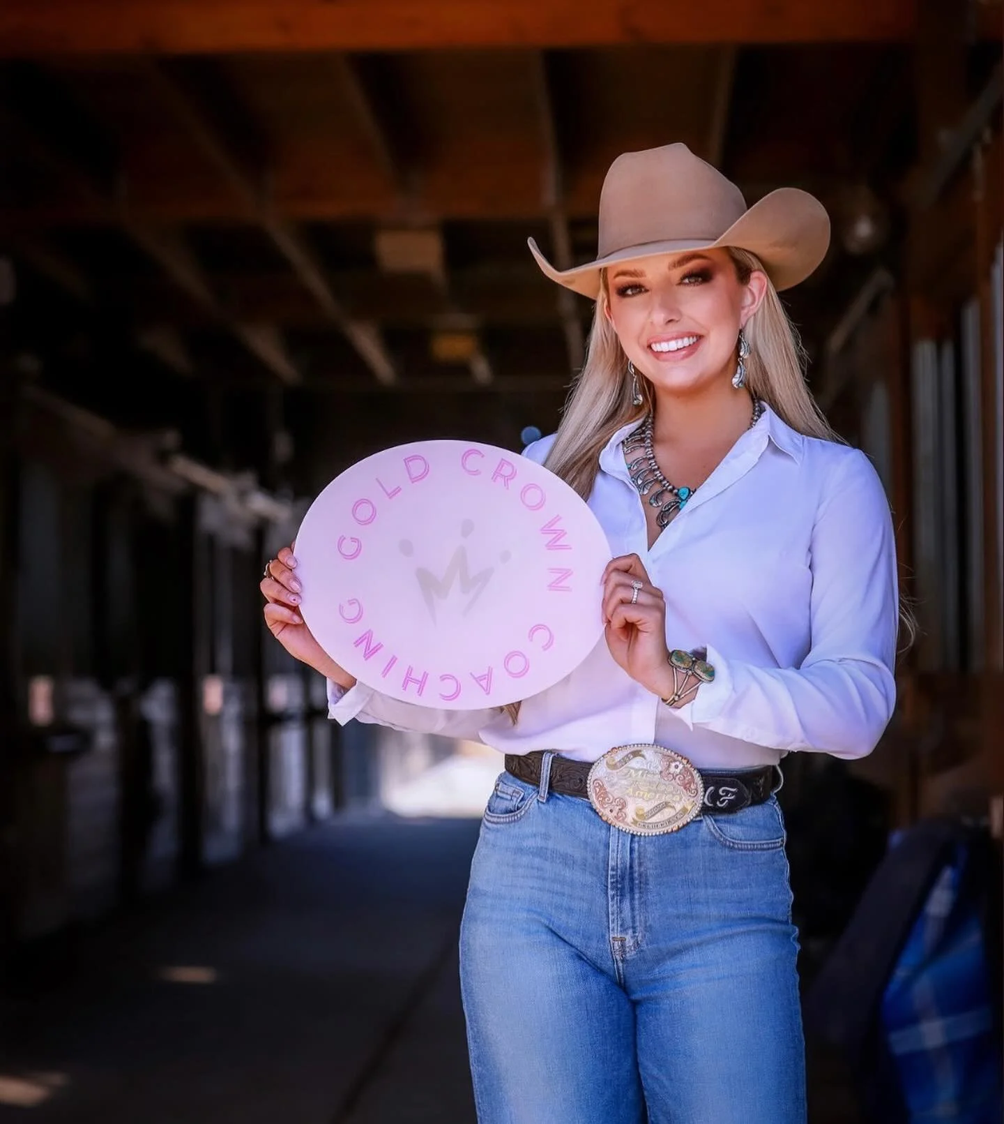 A woman dressed as a cowgirl, wearing a cowboy hat, white shirt, jeans, and jewelry, smiling and holding a round sign that reads 'GOLD CROWN CHOCHIN' in pink letters.
