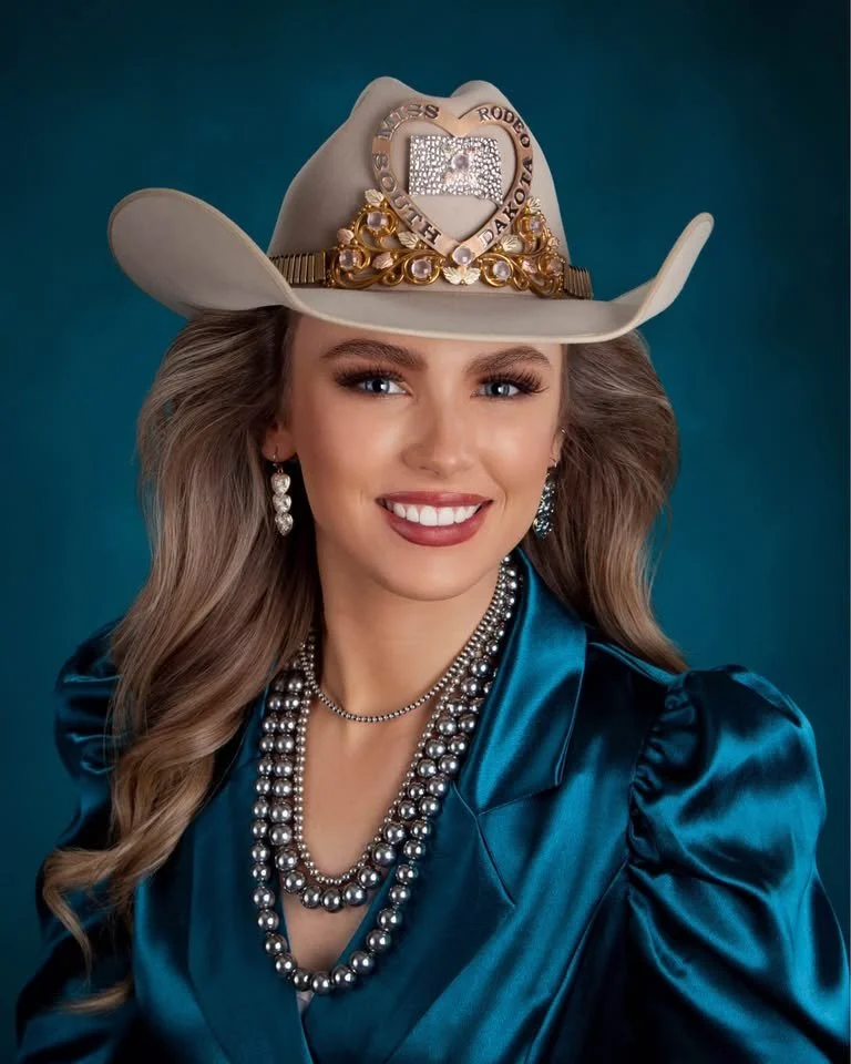 Young woman with long wavy hair wearing a cowboy hat decorated with gold and diamonds, a blue satin blouse, and layered pearl necklaces, smiling at the camera against a dark background.