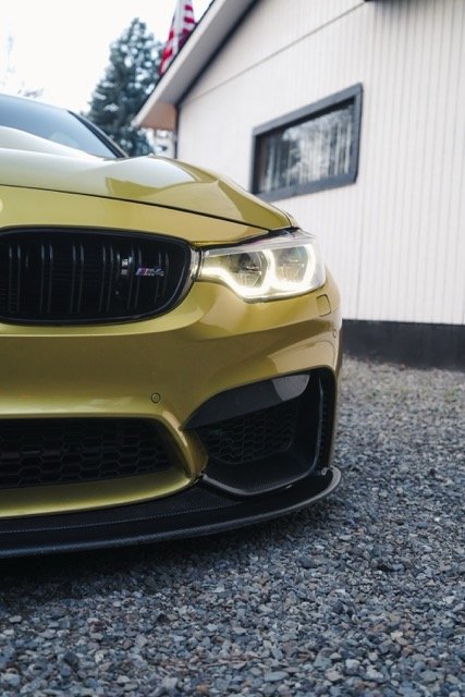 Yellow sporty car front view with headlight on, parked on gravel in front of a building.