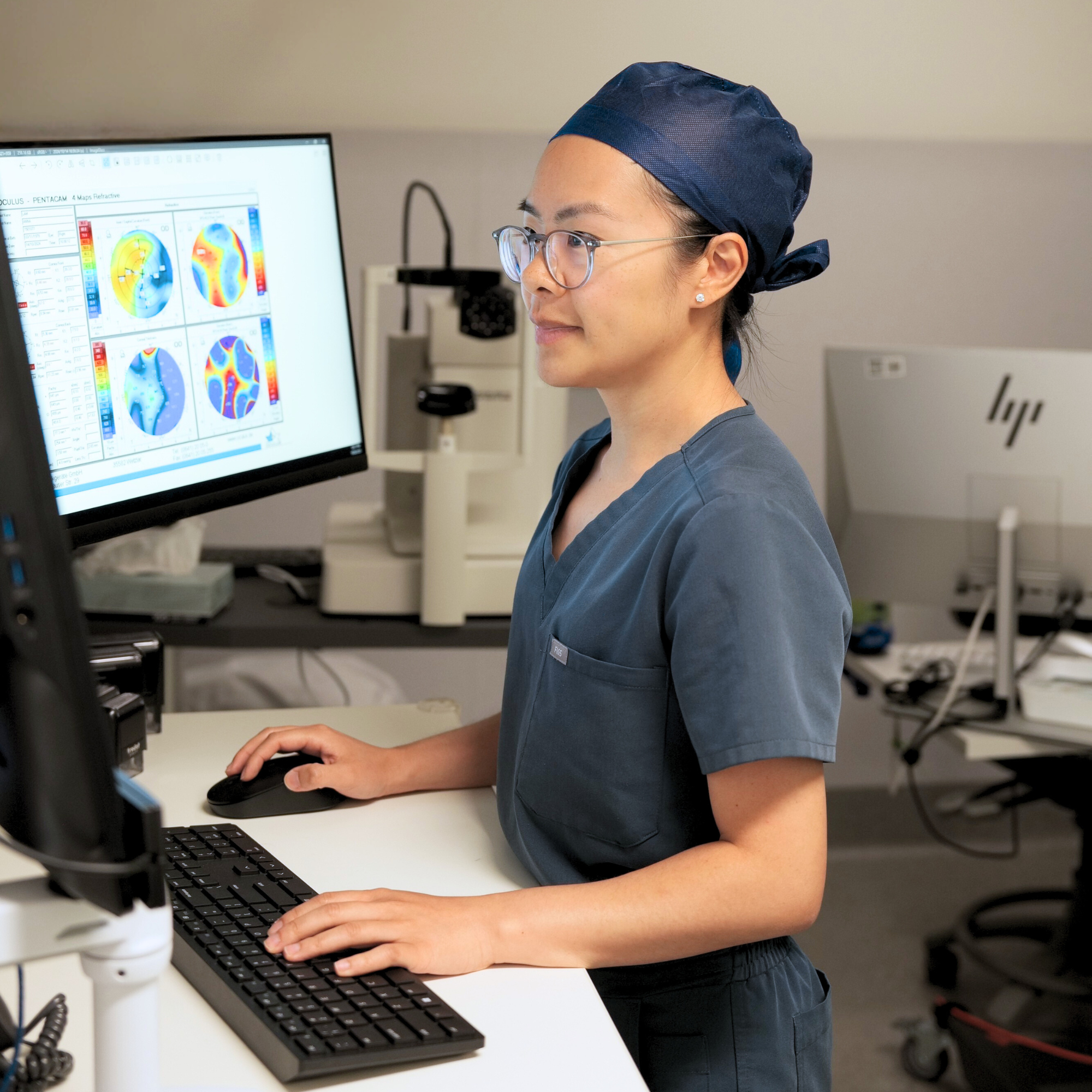 A medical professional wearing blue scrubs, a navy surgical cap, and glasses working at a computer station with a monitor displaying brain scan images.