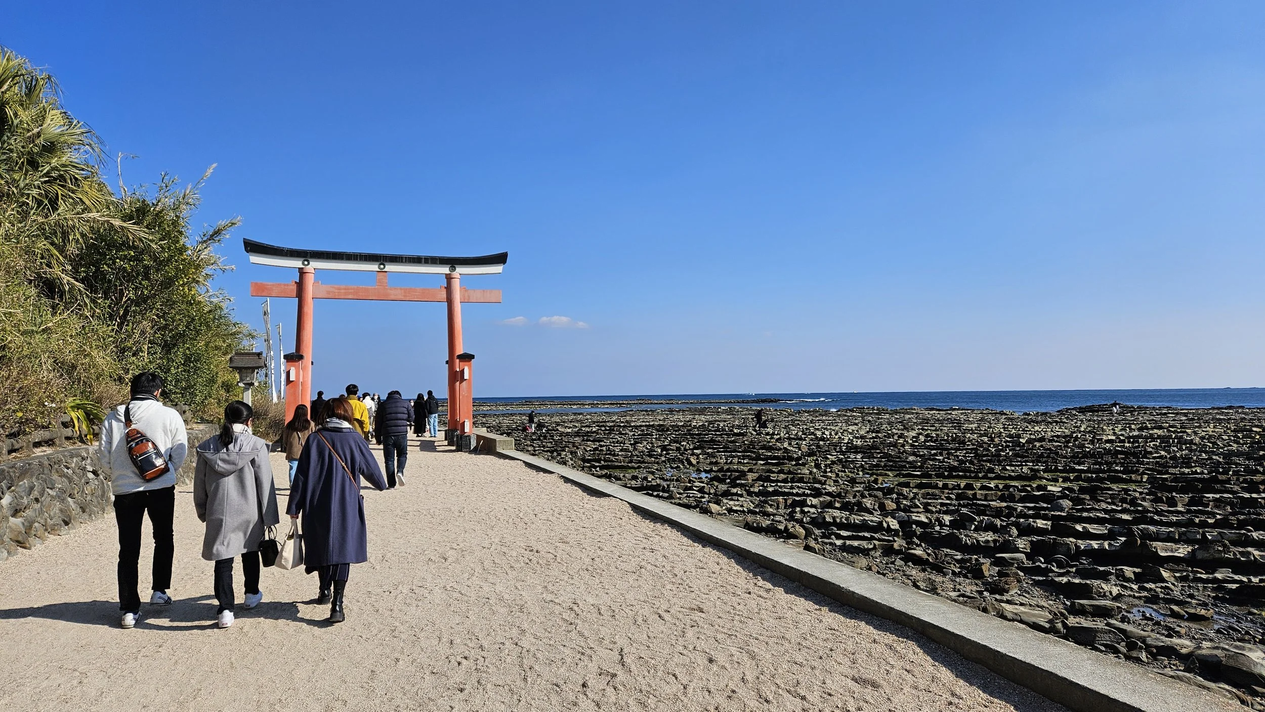 Tourists walking along a path past a large red torii gate towards the ocean on a sunny day.
