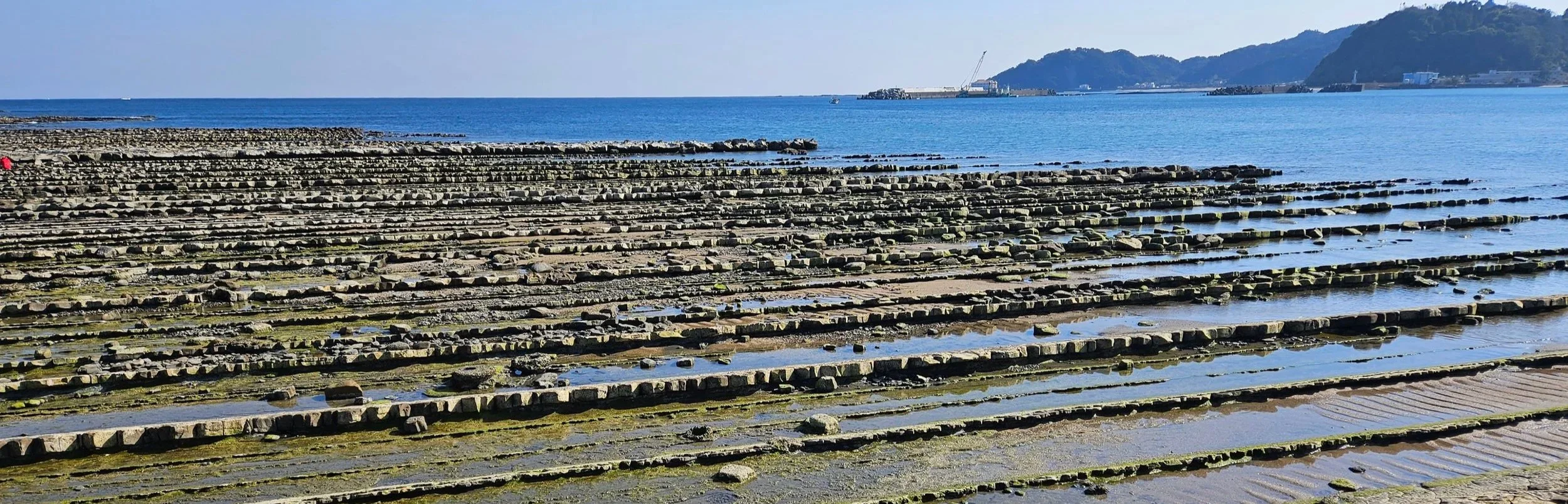 Coastal scene with a rocky shoreline featuring evenly spaced, flat stone formations extending into the water, with distant hills and a ship in the background.