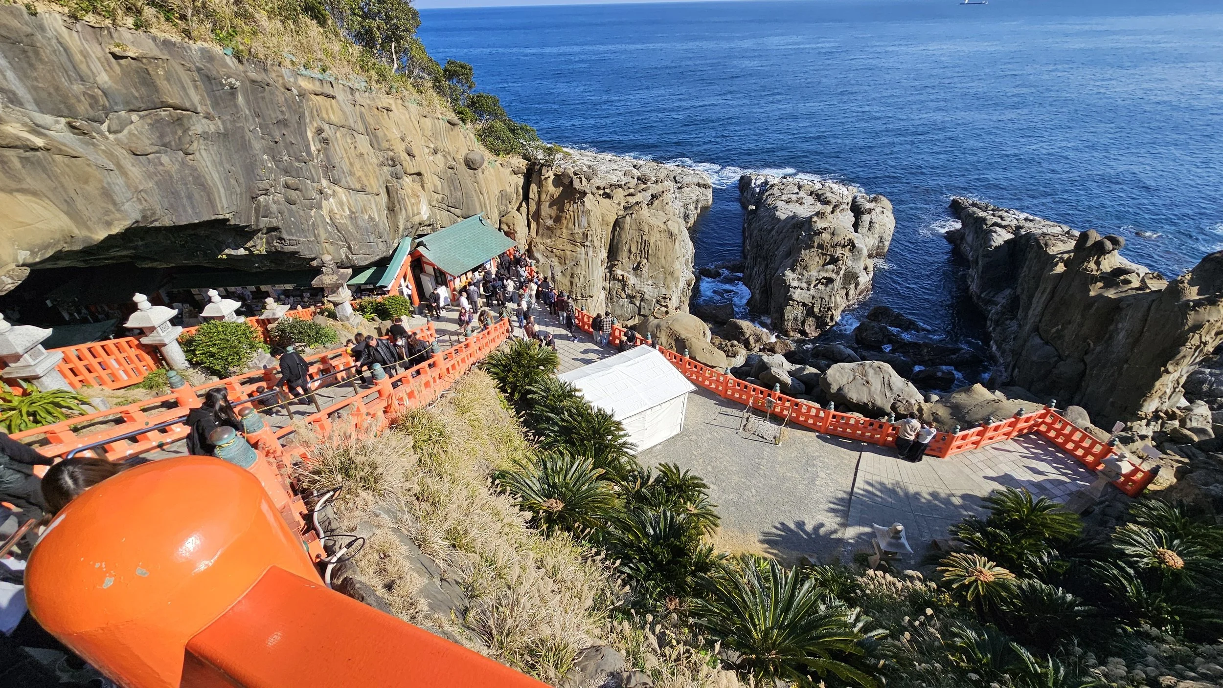 People walking on a path with red railings along rocky cliffs by the ocean, with clear blue water and sky in the background.