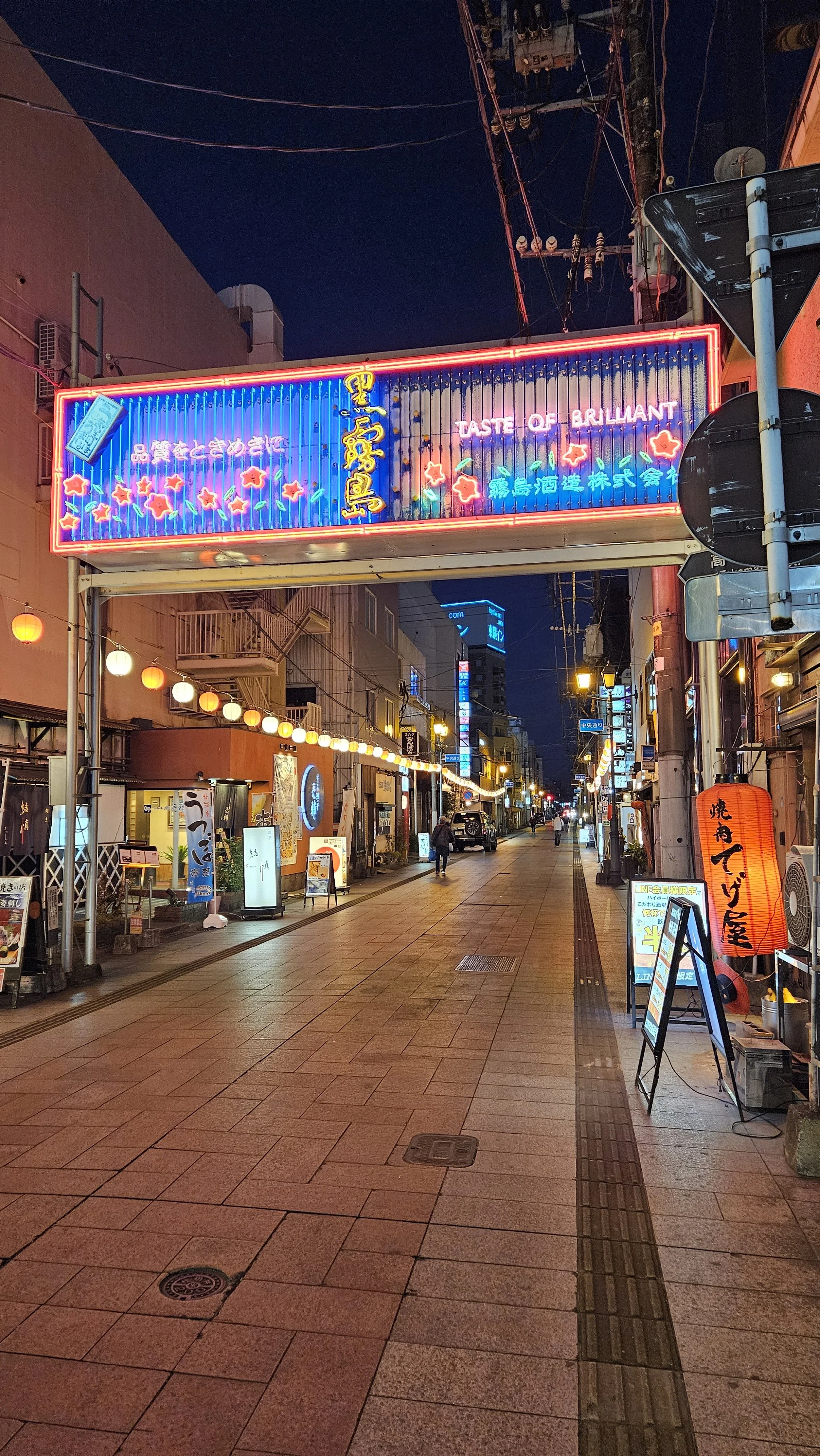A lively street scene at night with neon signs, lanterns, and shops, illuminated by colorful lights and strings of lanterns, in a busy area.