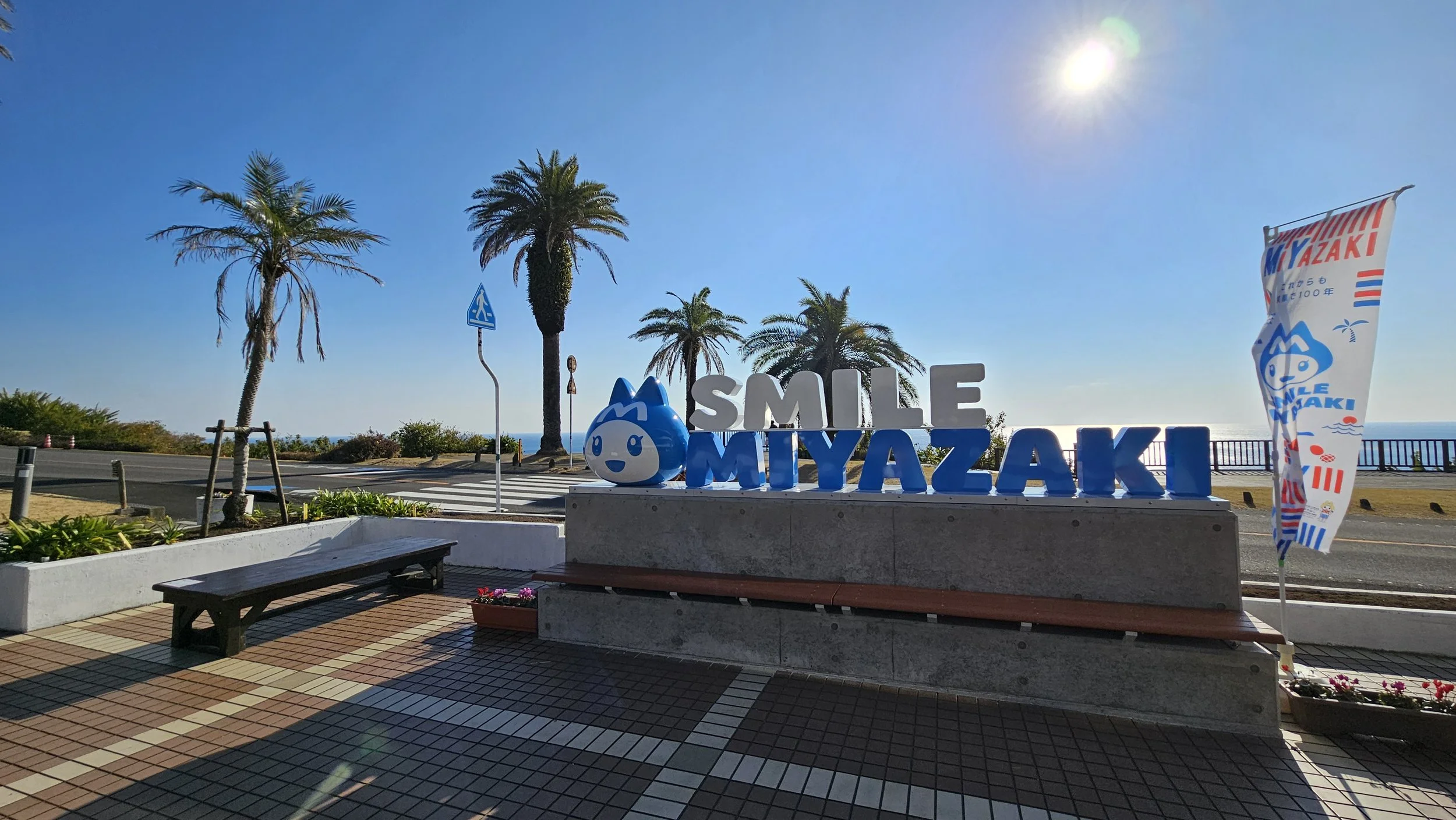 Sculpture of the words 'SMILE MIYAZAKI' with a smiling water droplet character and palm trees in the background along a seaside promenade.