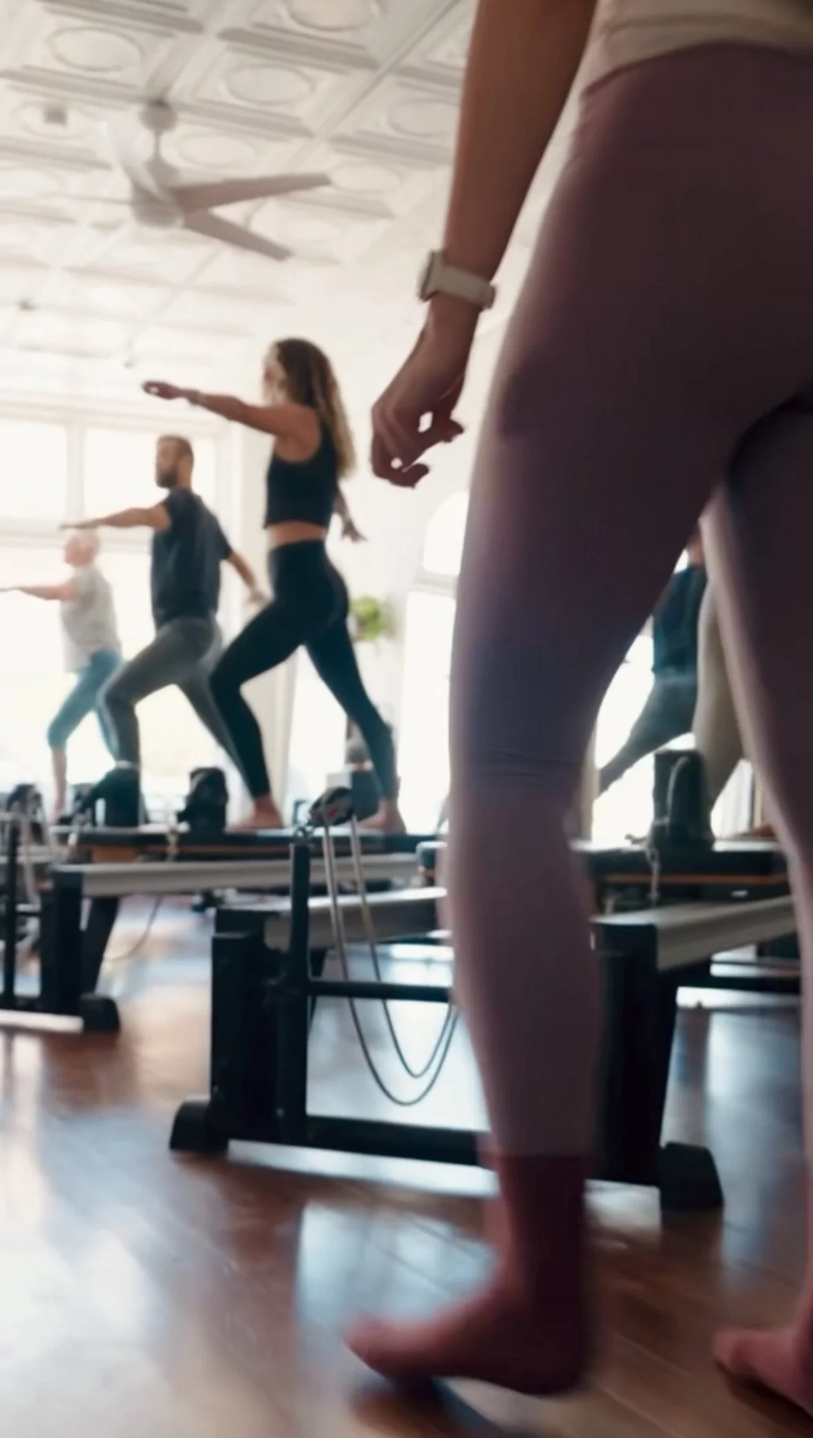 People participating in a Pilates or dance class in a bright, spacious room with wooden floors, some on reformer machines, with sunlight streaming through large windows.