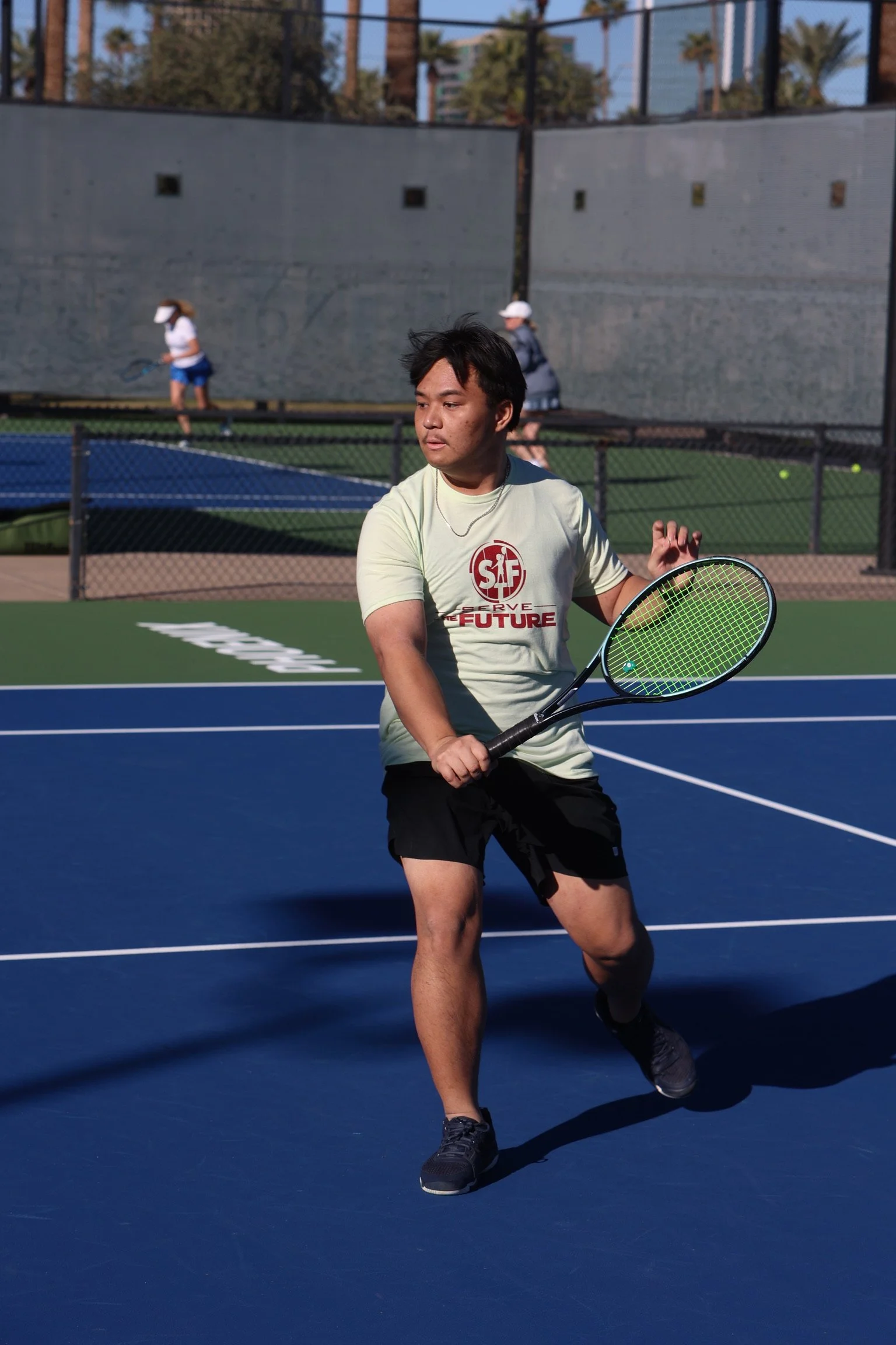 A man playing tennis on an outdoor court with a ladylike playing tennis in the background.