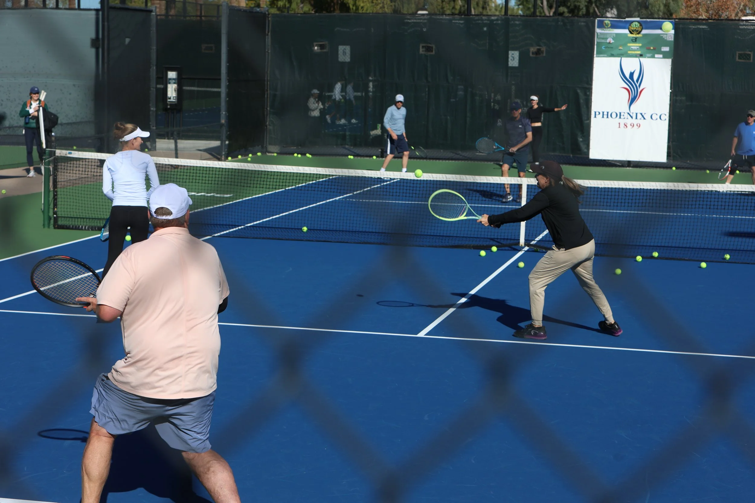 People playing pickleball on a blue court with a net, with tennis balls scattered around. The court has a sign that reads 'PHOENIX CC 1899' in the background.