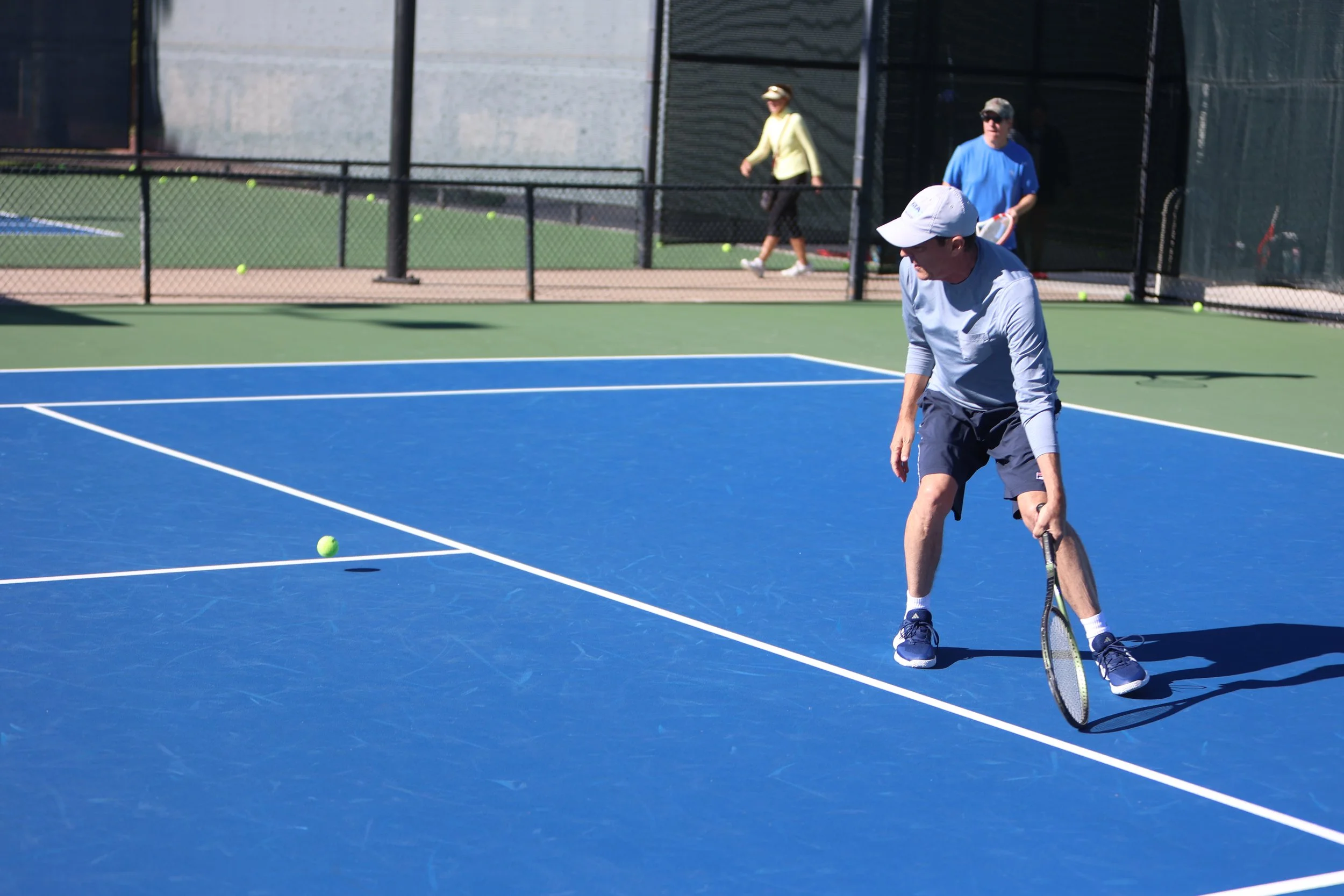 A man in a white cap and sunglasses preparing to hit a tennis ball on a blue tennis court with a green border, with two women in the background walking near a fence.