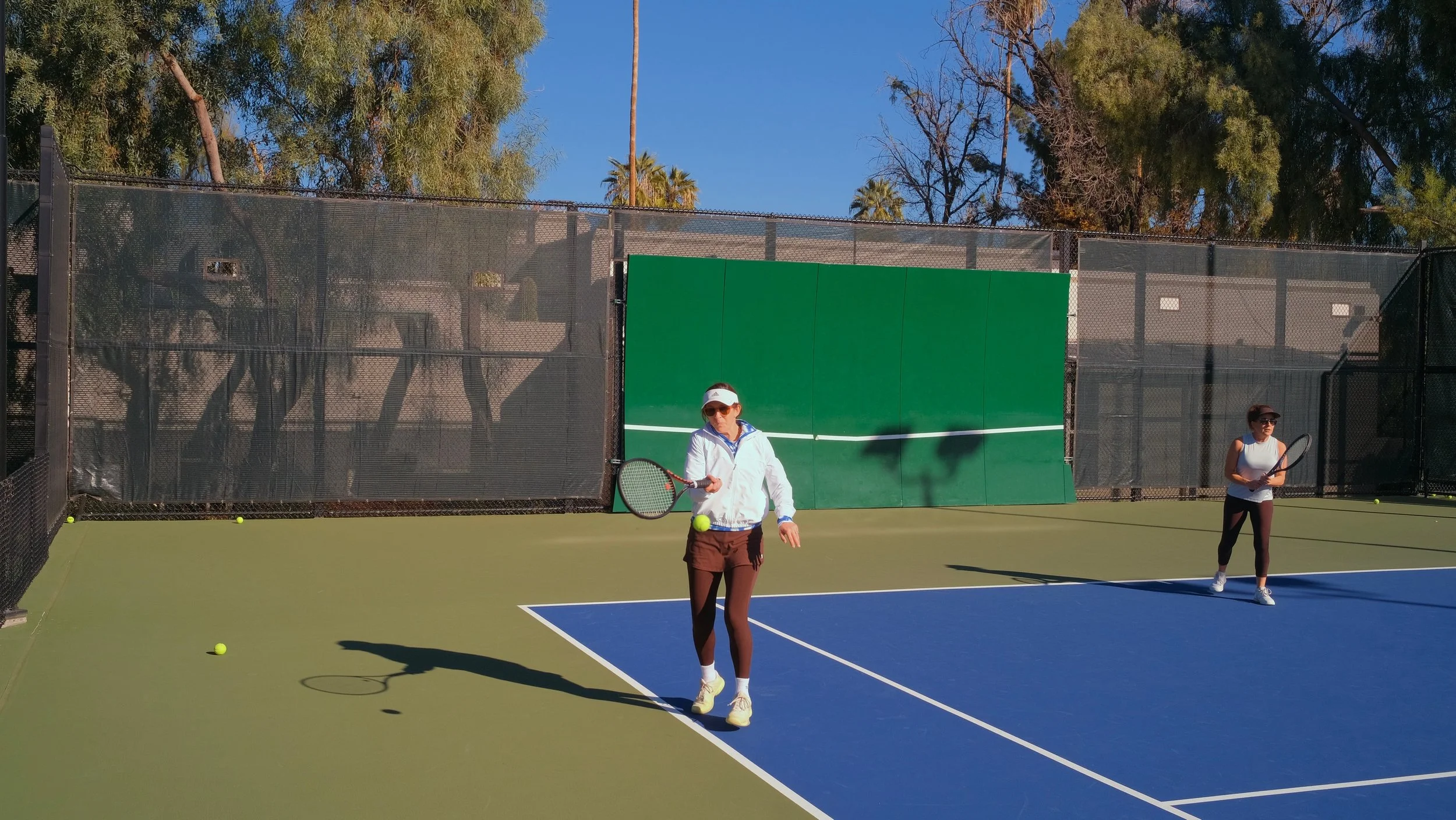 Two women playing tennis on an outdoor court with green and blue surfaces. One woman is in the foreground preparing to hit a tennis ball with her racket, while the other stands further back, holding a racket. The scene is sunny, with trees and a clea
