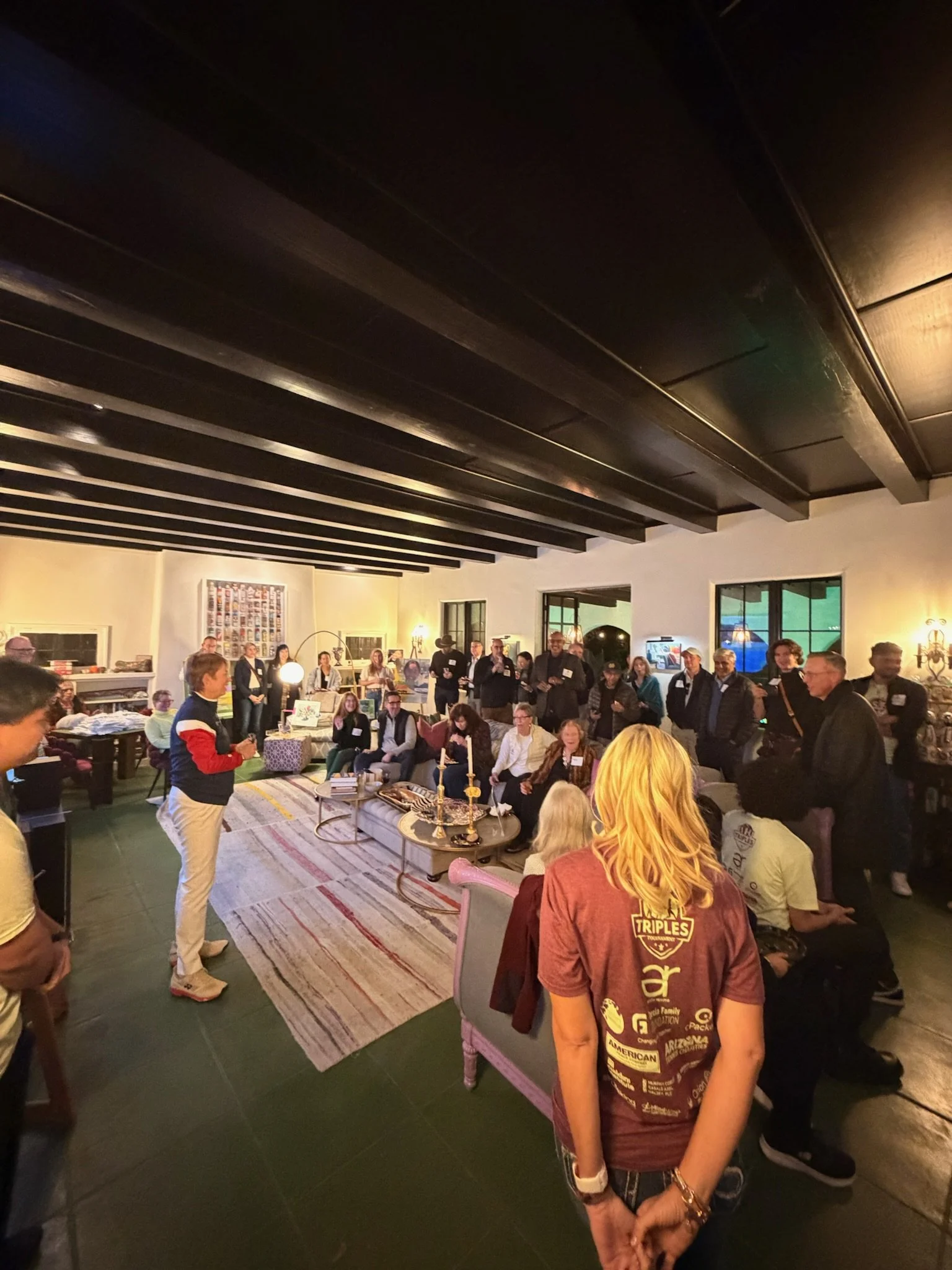 A group of people gathered in a cozy living room-like setting with modern decor, many sitting on couches and some standing, listening to a woman speak at the front. The room has a dark ceiling with wooden beams and large windows, warm lighting, and a