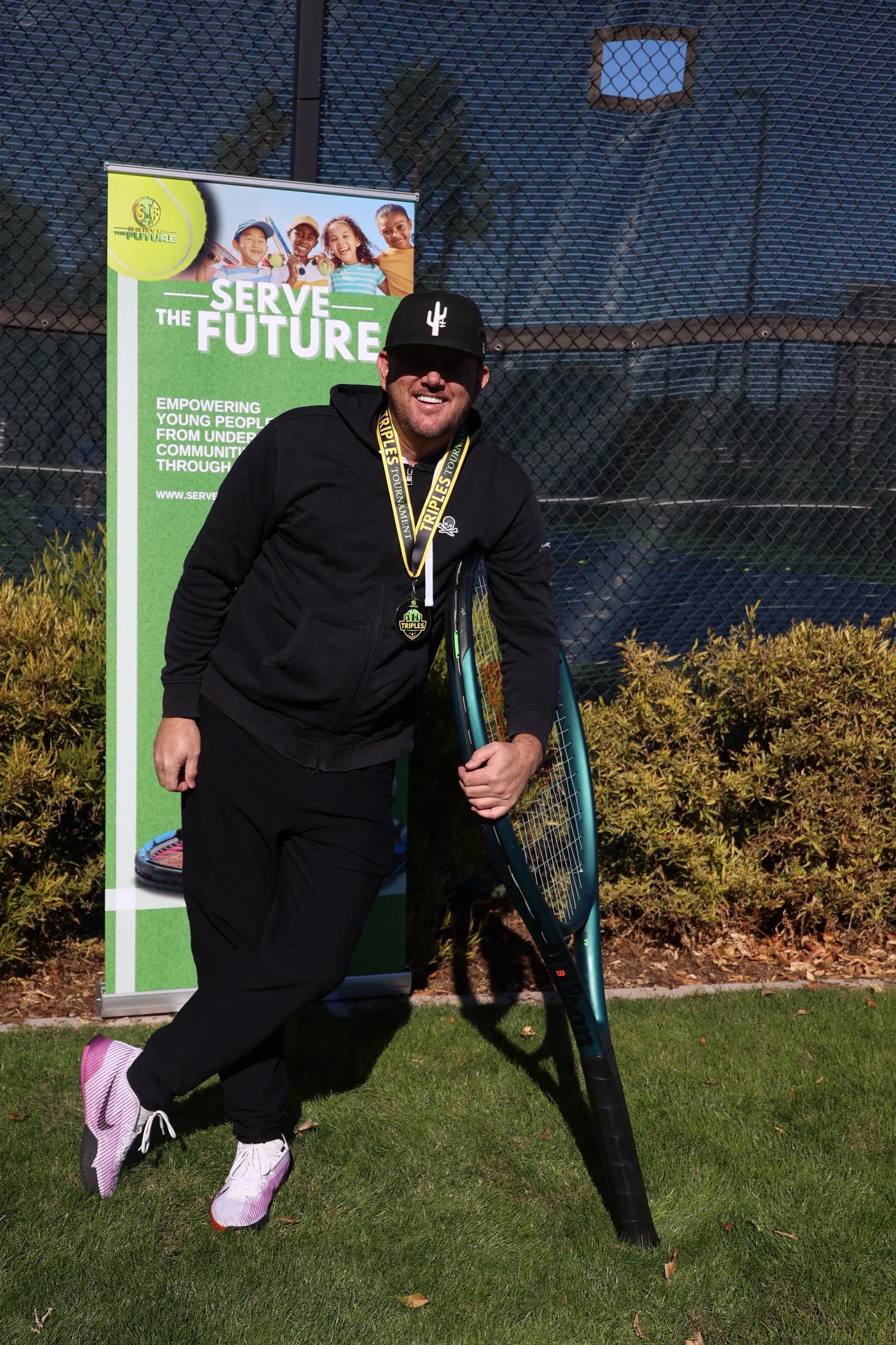 A man smiling and holding a tennis racket and pickleball paddle, standing in front of a donation or awareness sign for Serve the Future, outdoors on a grassy area with bushes and a fence in the background.