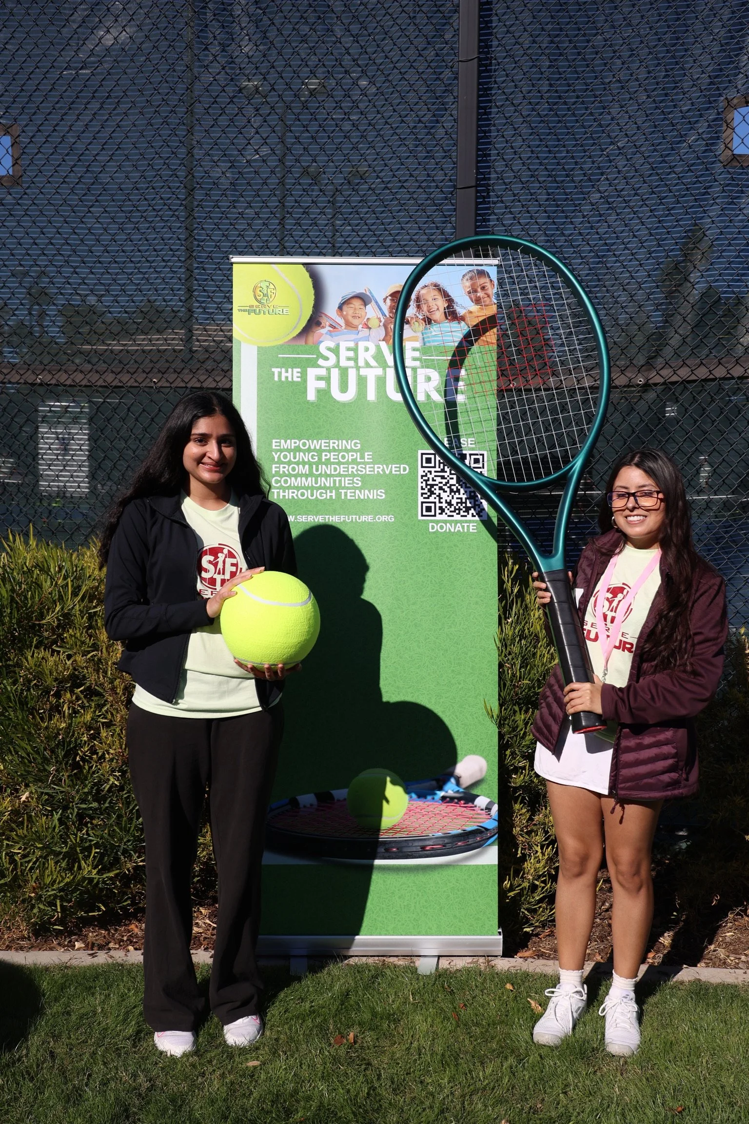 Two young women stand outdoors on a tennis court, holding tennis equipment and smiling. One holds a large tennis ball, and the other holds a tennis racket. Behind them is a green banner with the text 'Serve the Future' and a QR code, with a tennis co