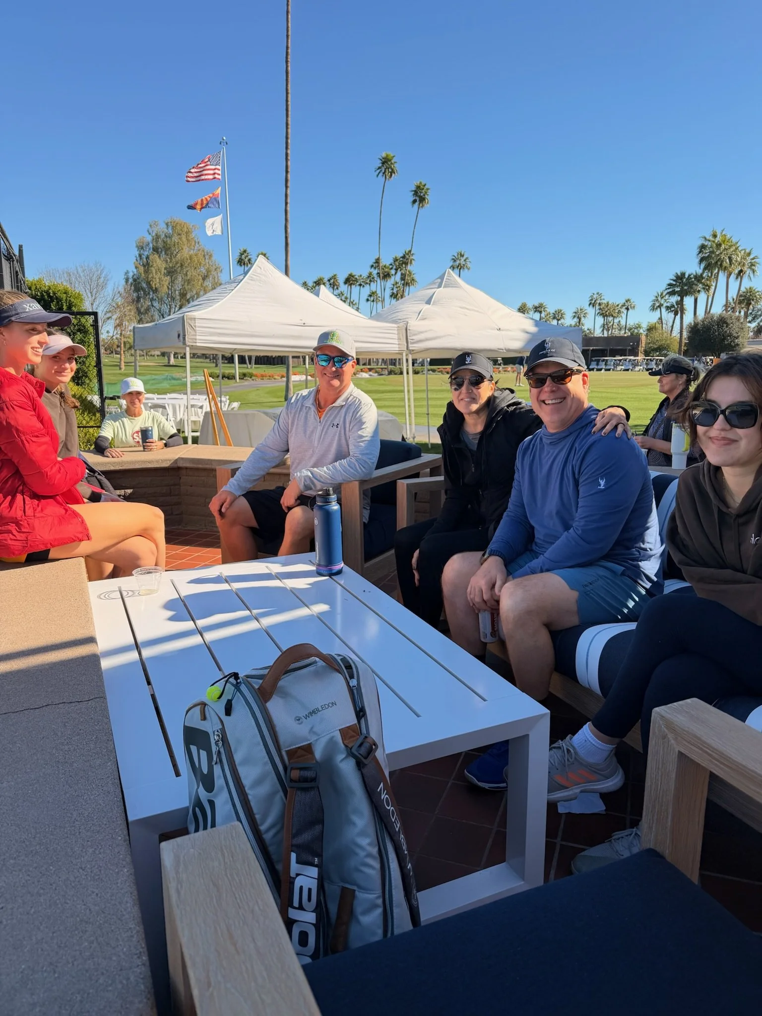 A group of people sitting at a table outdoors at a golf course, smiling and enjoying the sunny day with palm trees and flags in the background.