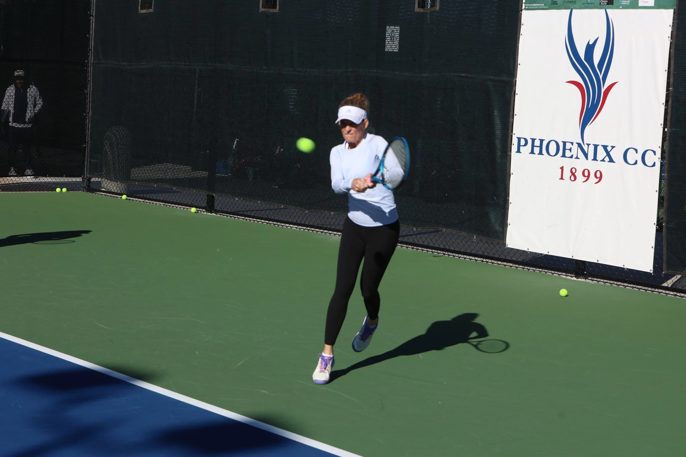 A woman playing tennis on a green outdoor court, preparing to hit a tennis ball with a racket. She is wearing a white long-sleeve shirt, black leggings, sneakers, and a white visor. There are tennis balls on the court and a banner with the logo and n