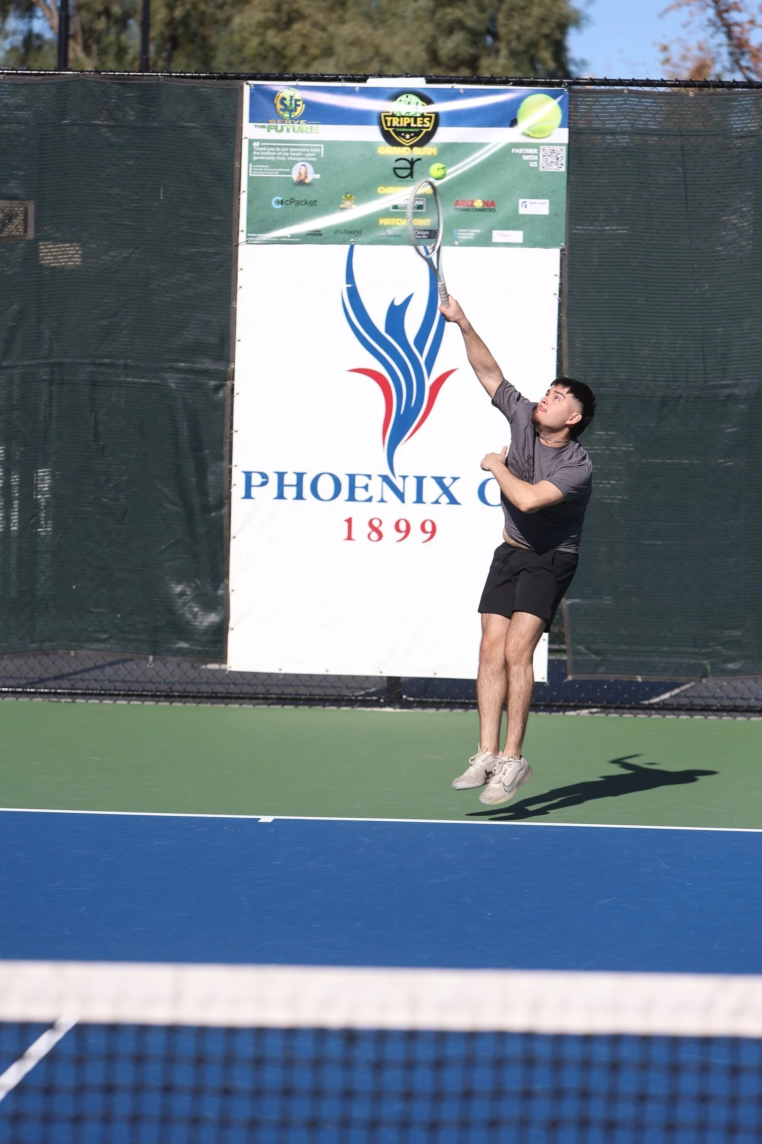 A man jumping to hit a tennis ball with his racket on an outdoor tennis court, with a sign in the background displaying 'Phoenix 1899' and a logo.