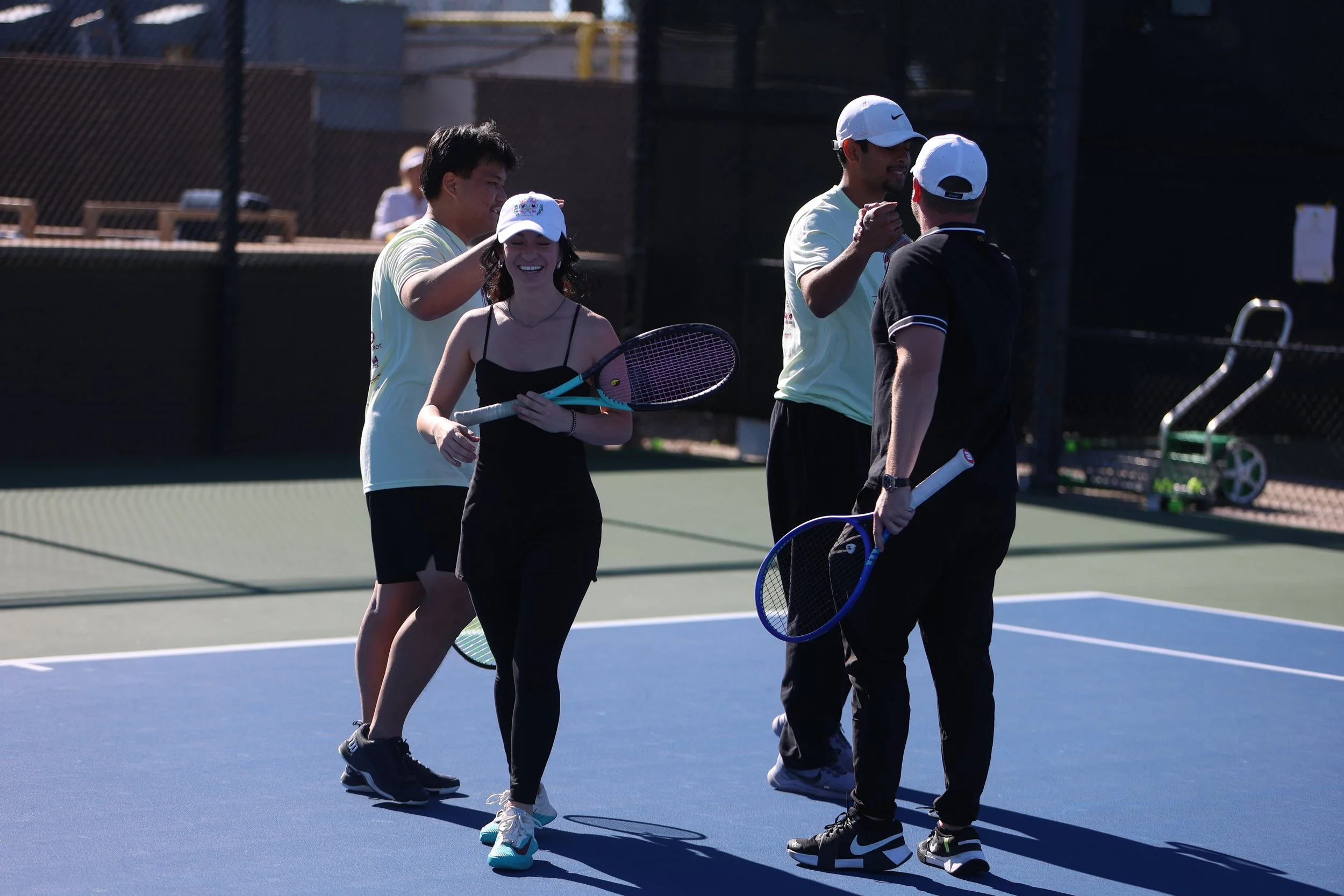Group of four tennis players celebrating on a tennis court, two men and one woman smiling, holding tennis rackets, with one man giving a high five.