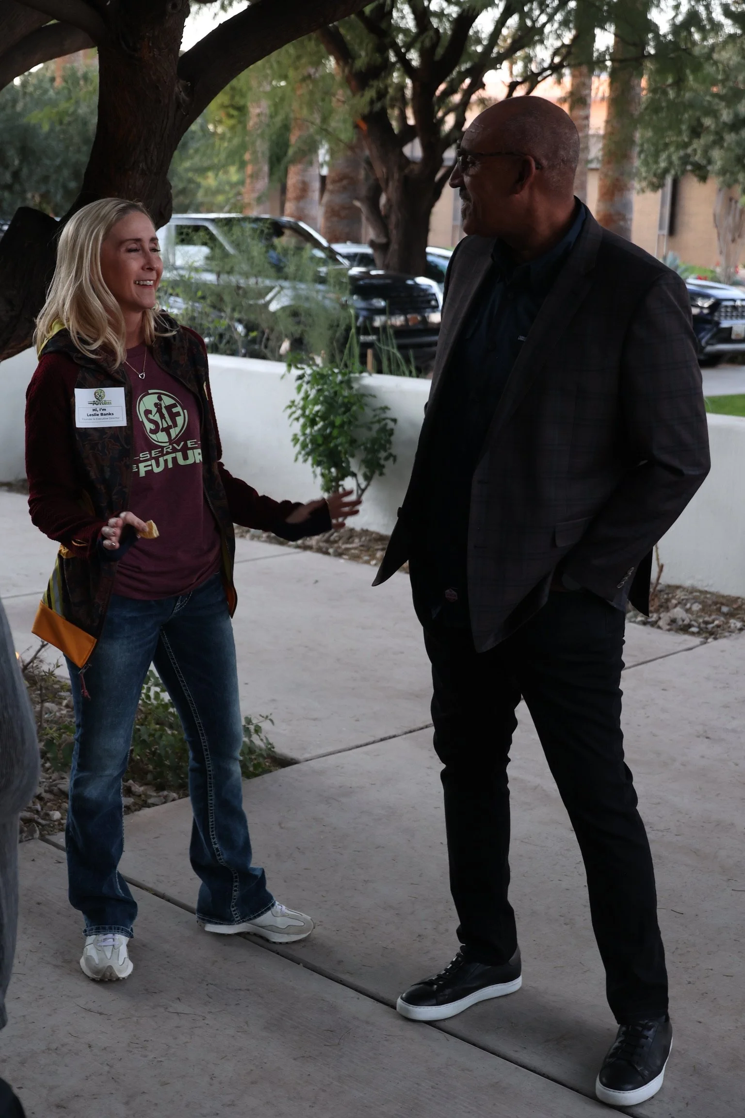 A woman and man are laughing and talking outdoors under a large tree, with parked cars and a building in the background.